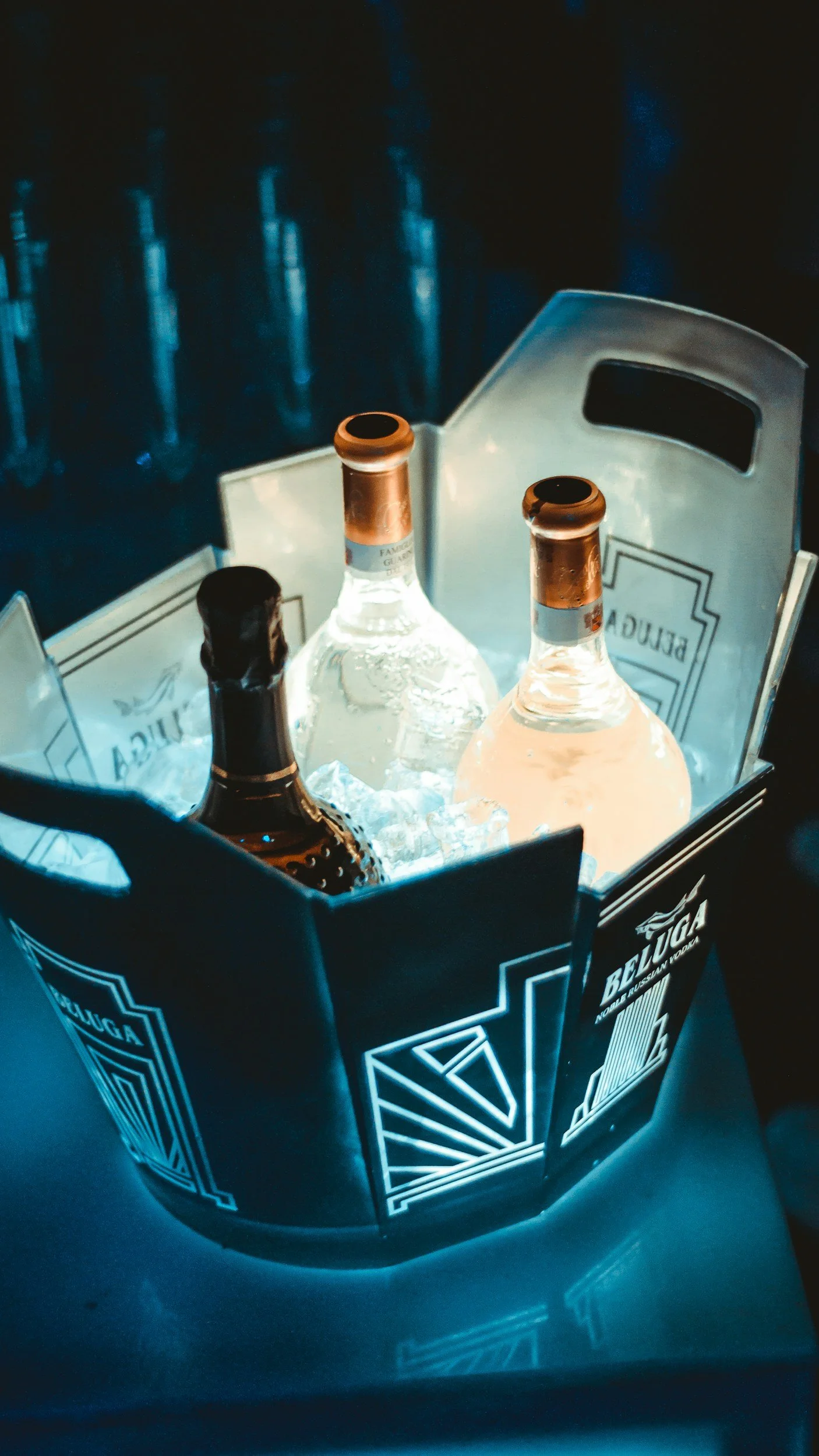 A metal ice bucket containing three bottles of alcohol, two clear and one dark, sitting on a surface with an ambient blue lighting.