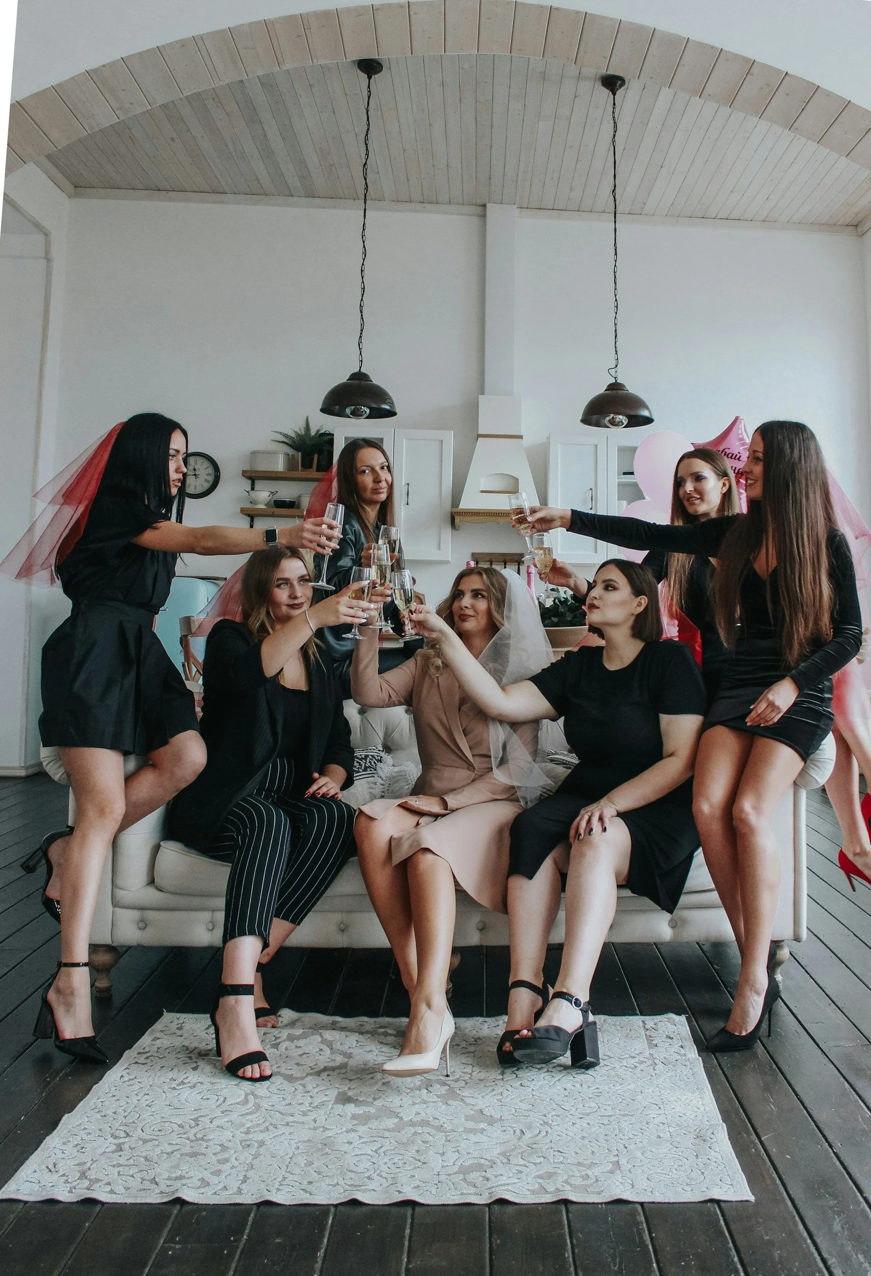 Group of women sitting and standing on a sofa, celebrating a bridal shower with champagne glasses, some wearing veils and pink party accessories.