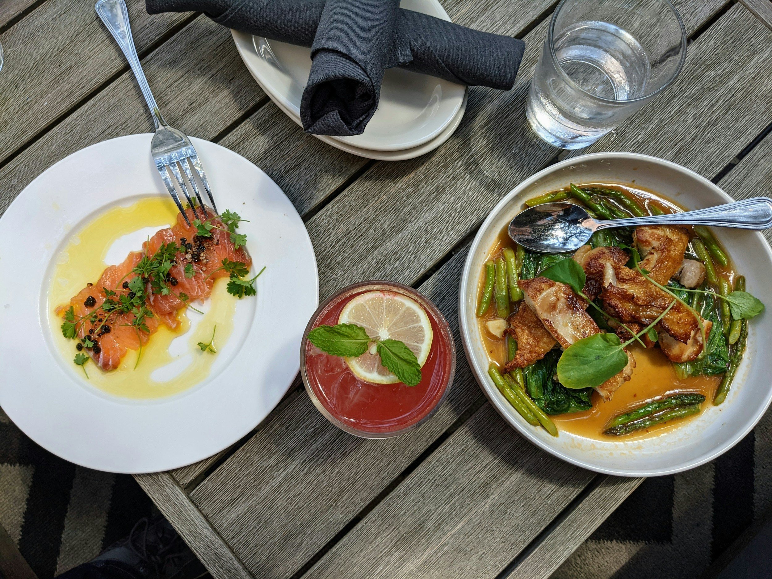 A table with a plate of sliced salmon topped with herbs and black pepper, a bowl of chicken with green beans and greens in a yellow broth, a glass of water, a glass of pink-colored beverage with lemon and mint, and a set of black napkins and utensils.