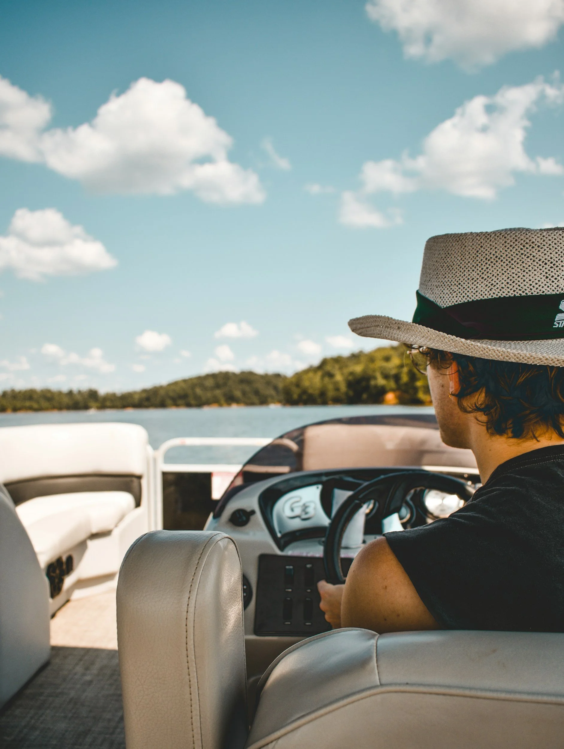 A person with curly hair wearing a straw hat and black sleeveless shirt, sitting at the helm of a boat, with a scenic lake and tree-covered shoreline under a partly cloudy sky in the background.
