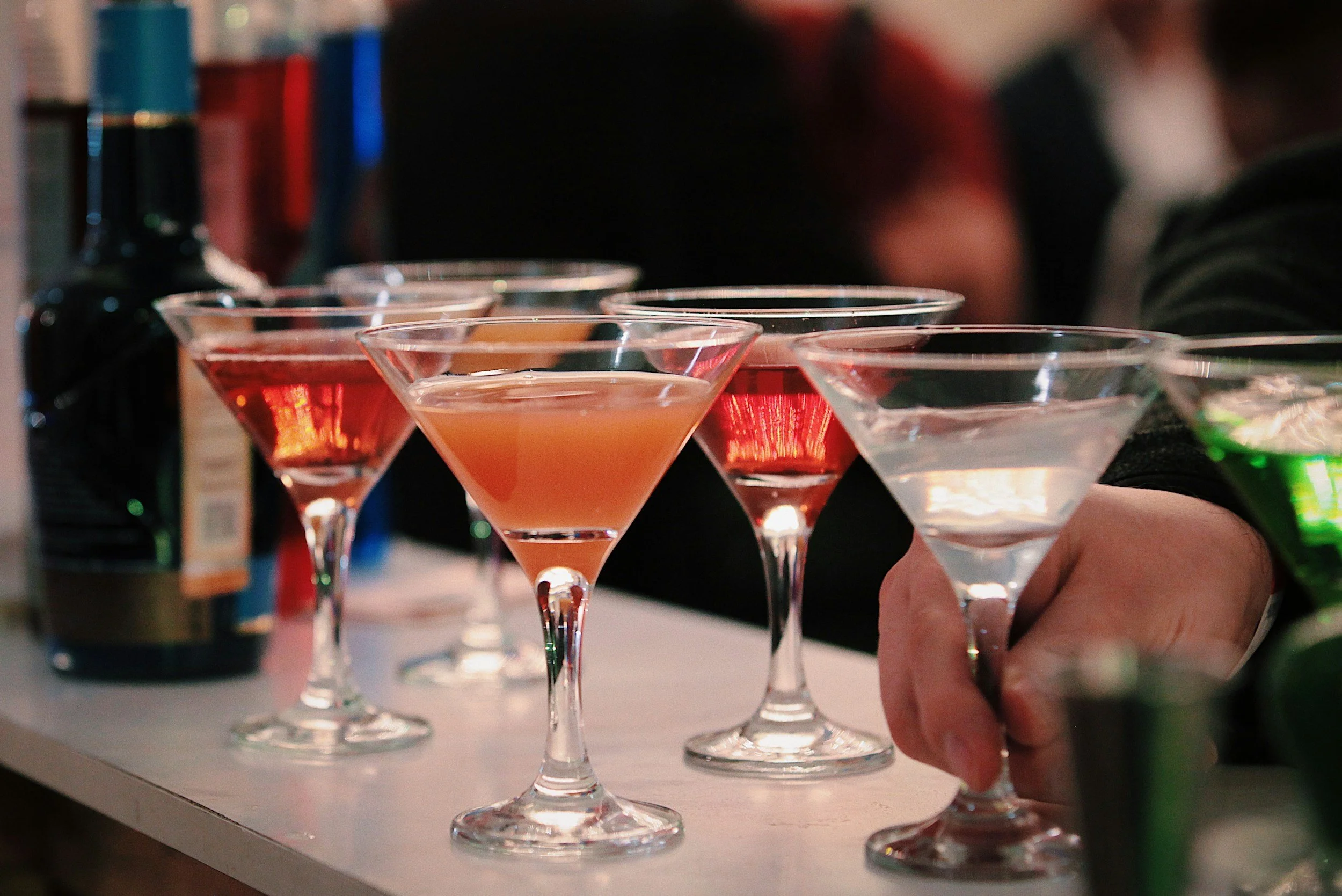 Multiple colorful cocktails in martini glasses set on a bar counter with a hand holding one glass, blurred background of people, and bottles in a bar setting.