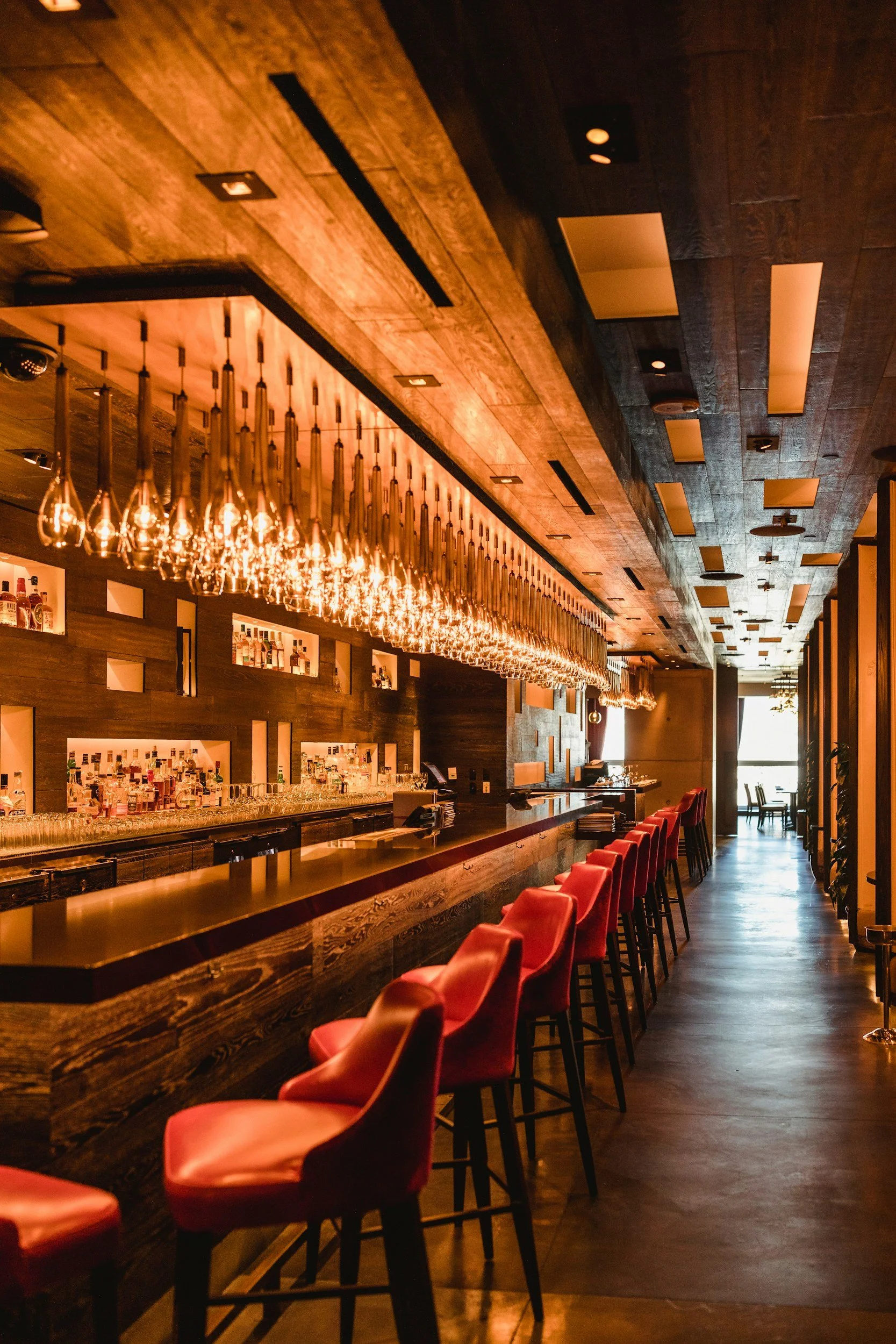 Empty bar with a row of red chairs, wooden counter, shelves of liquor bottles, and hanging wine glasses, warm lighting, and window with natural light.