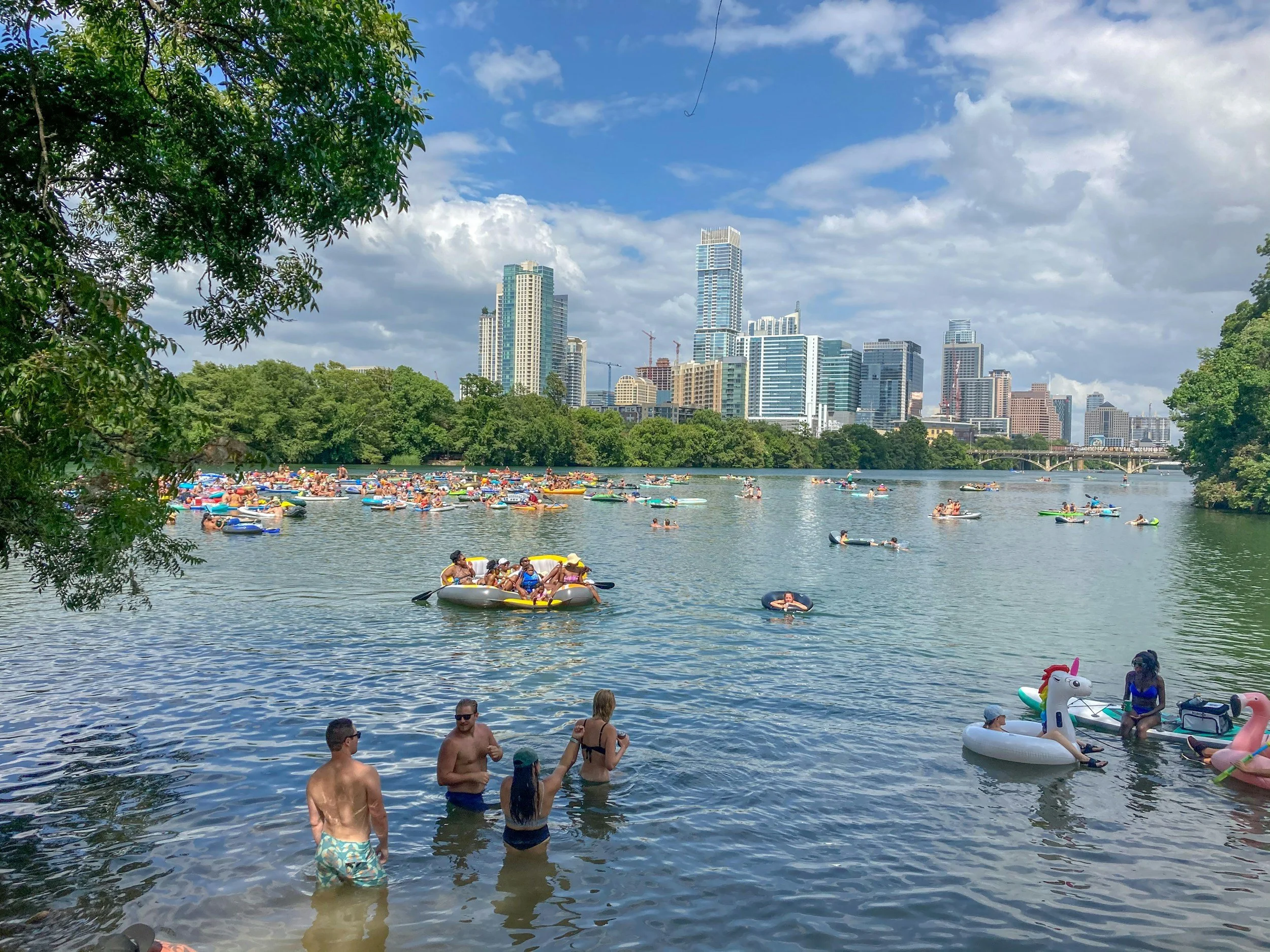 People swimming and floating on inflatable tubes in a city lake with a skyline of tall buildings in the background on a sunny day.