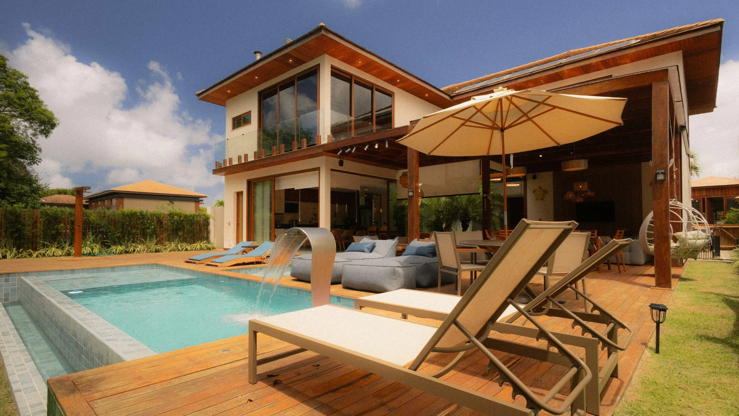 Modern two-story house with a swimming pool in the backyard. The pool has a waterfall feature, surrounded by wooden decking with lounge chairs, umbrella, and outdoor furniture. The house has large glass windows and a covered patio area.
