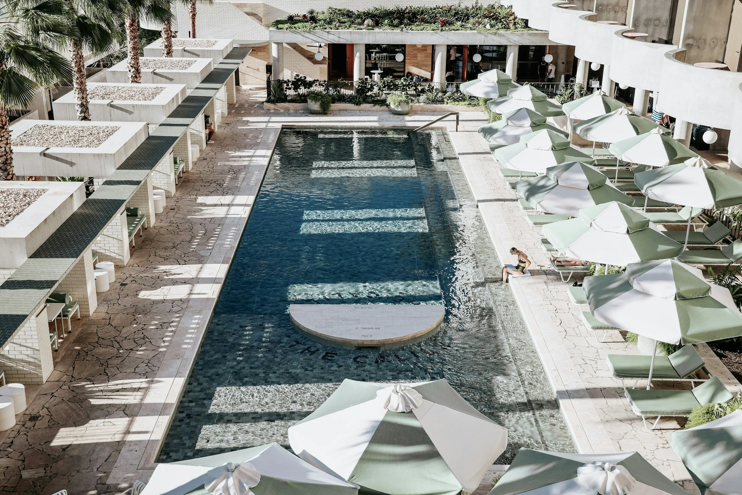 An outdoor hotel pool area with a person sitting on the poolside and white umbrellas shading lounge chairs.
