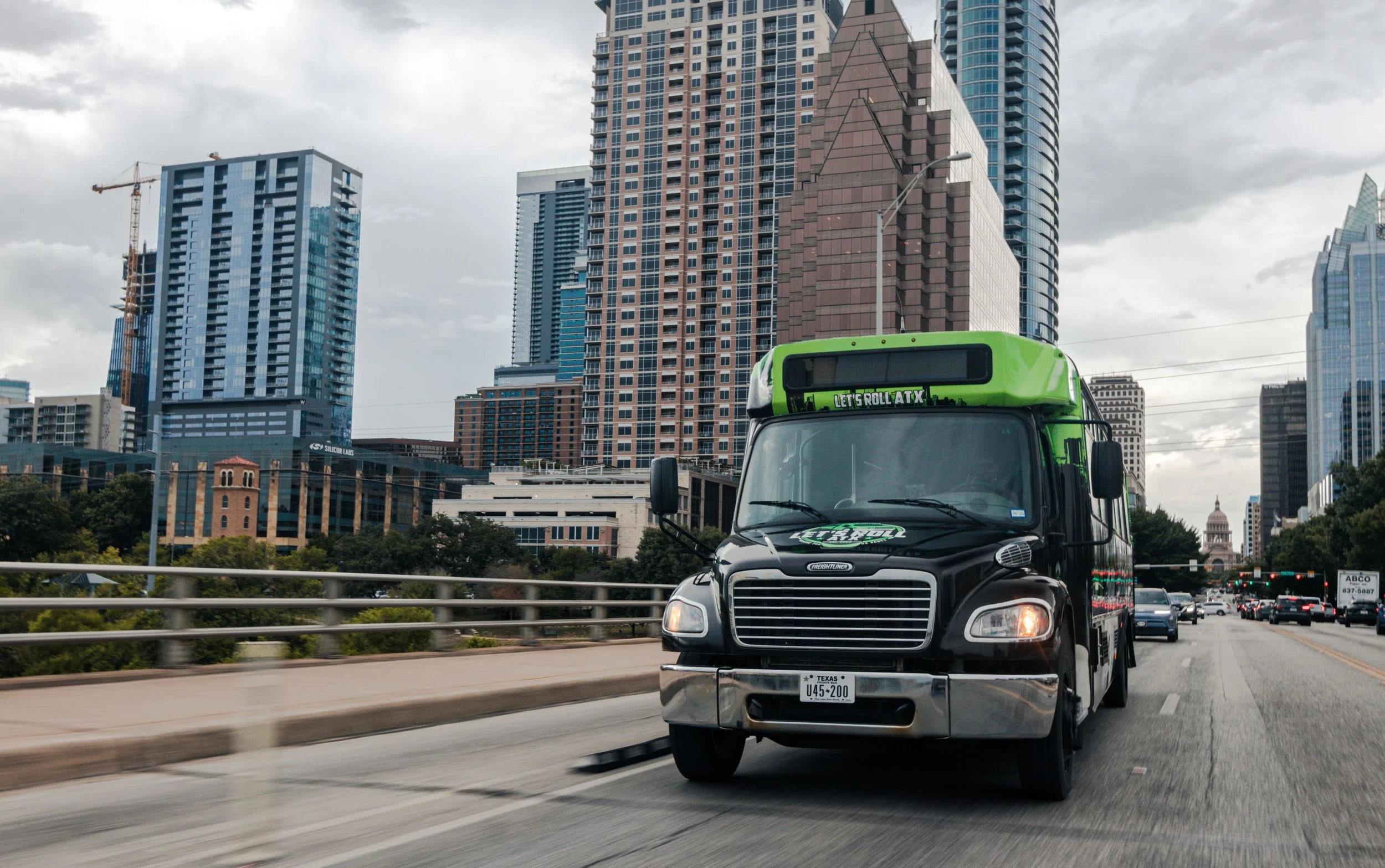 A black and green food truck with the text 'Let's Roll ATX' on the front, driving on a city street with tall modern buildings in the background and a cloudy sky.