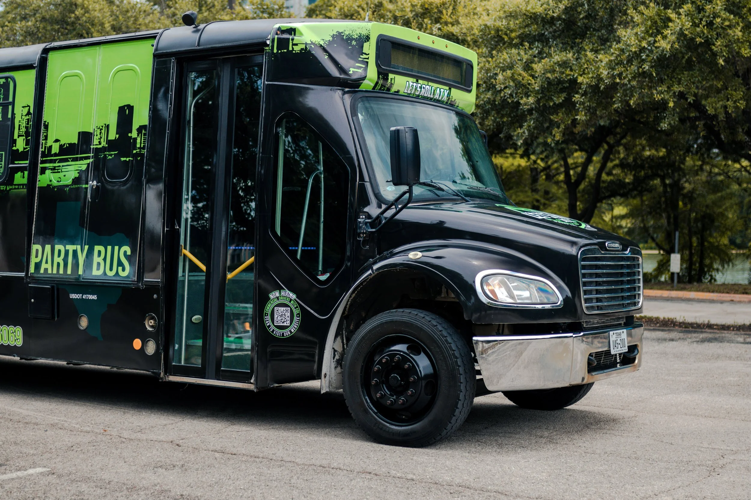 A black party bus with green accents and cityscape graphics on the side, parked on a street with trees in the background.