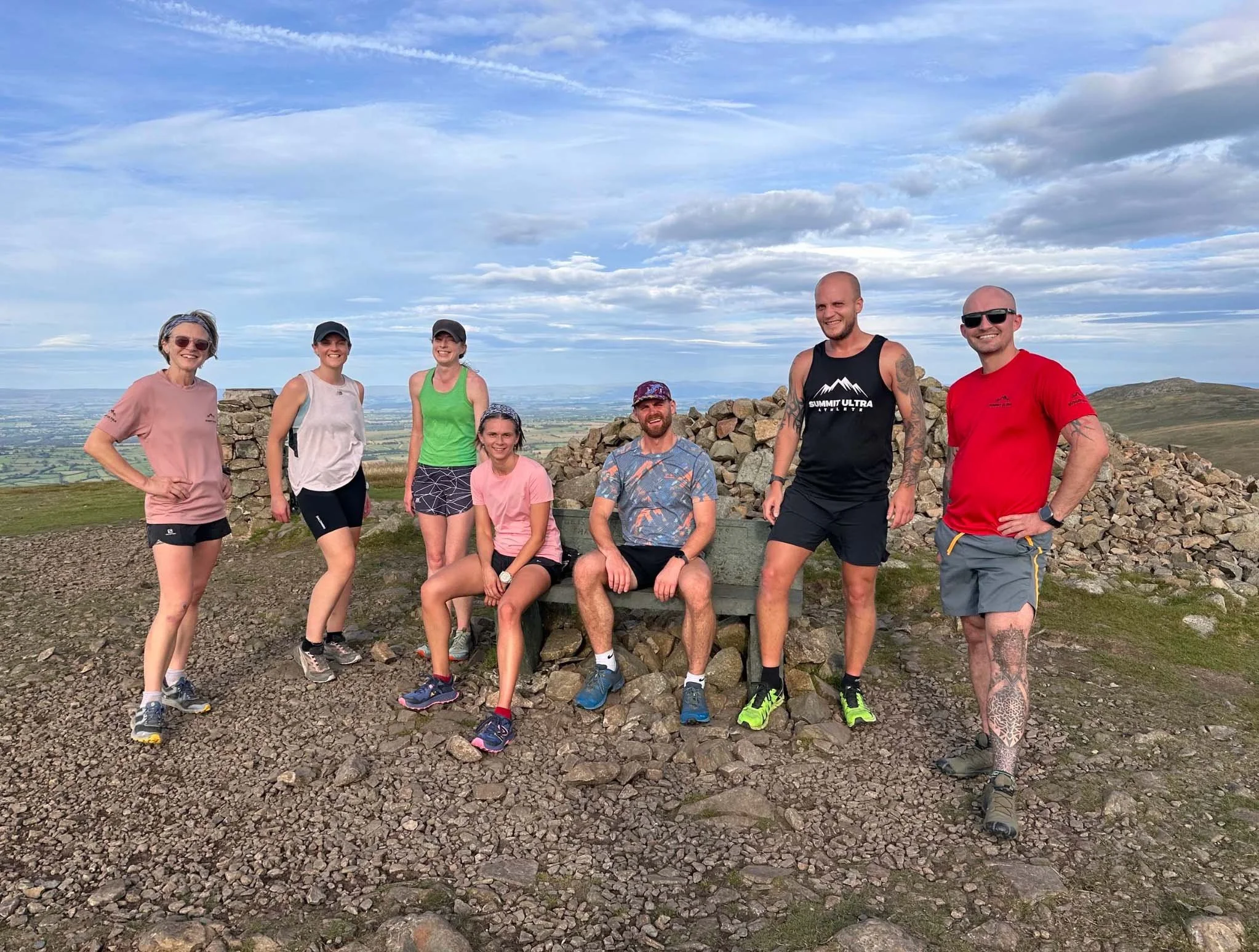Group of eight people hiking on a mountain summit, some sitting on a bench, with a vast landscape and cloudy sky in the background.