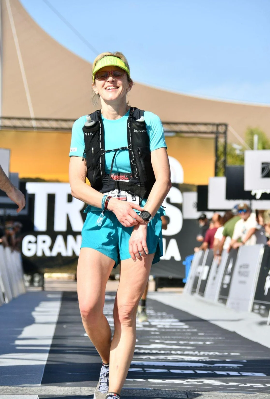A woman in running gear crossing a finish line at a marathon, smiling with sunlight and spectators in the background.