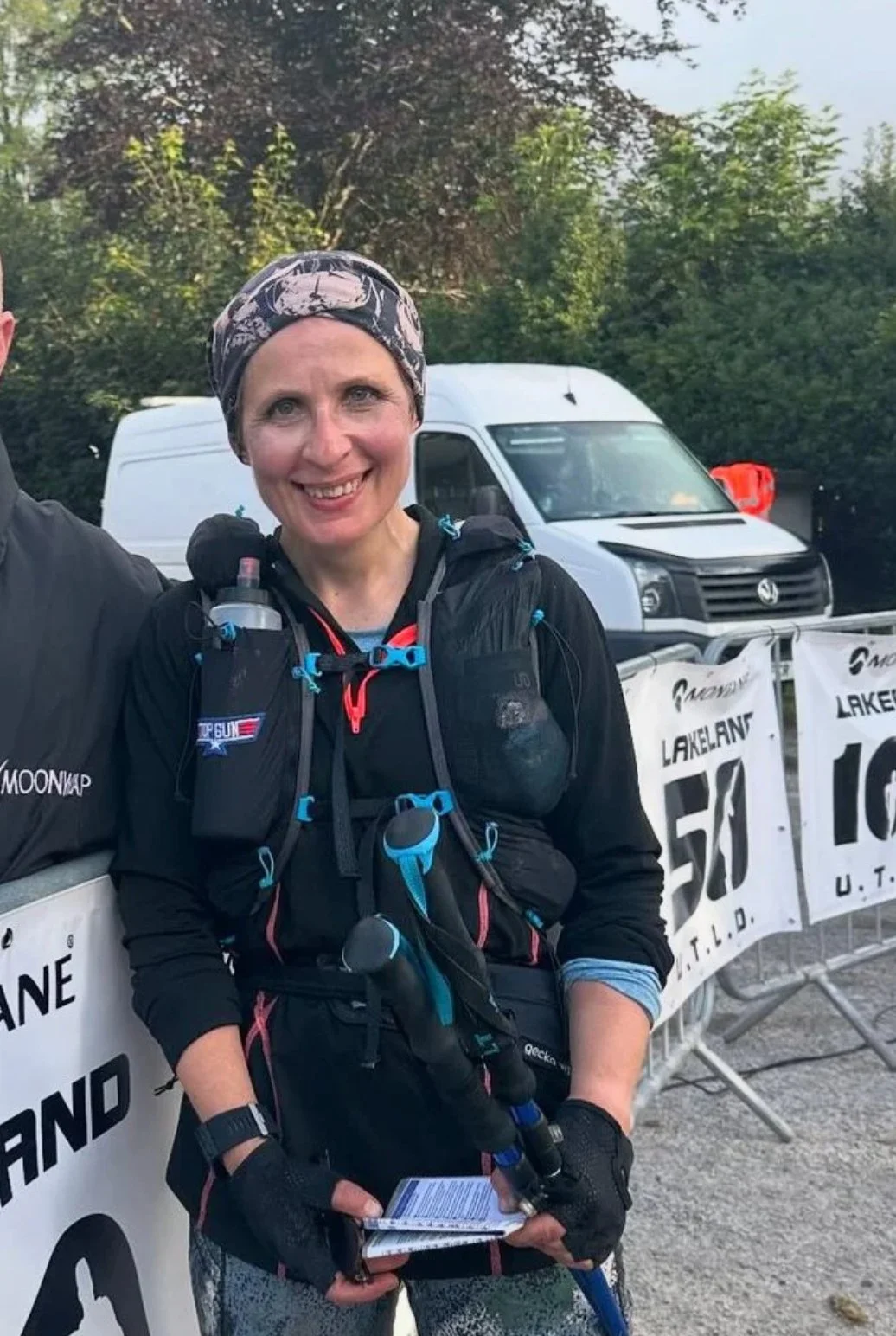 Female runner in athletic gear, smiling, holding a race number, standing outdoors near a white van and banner.