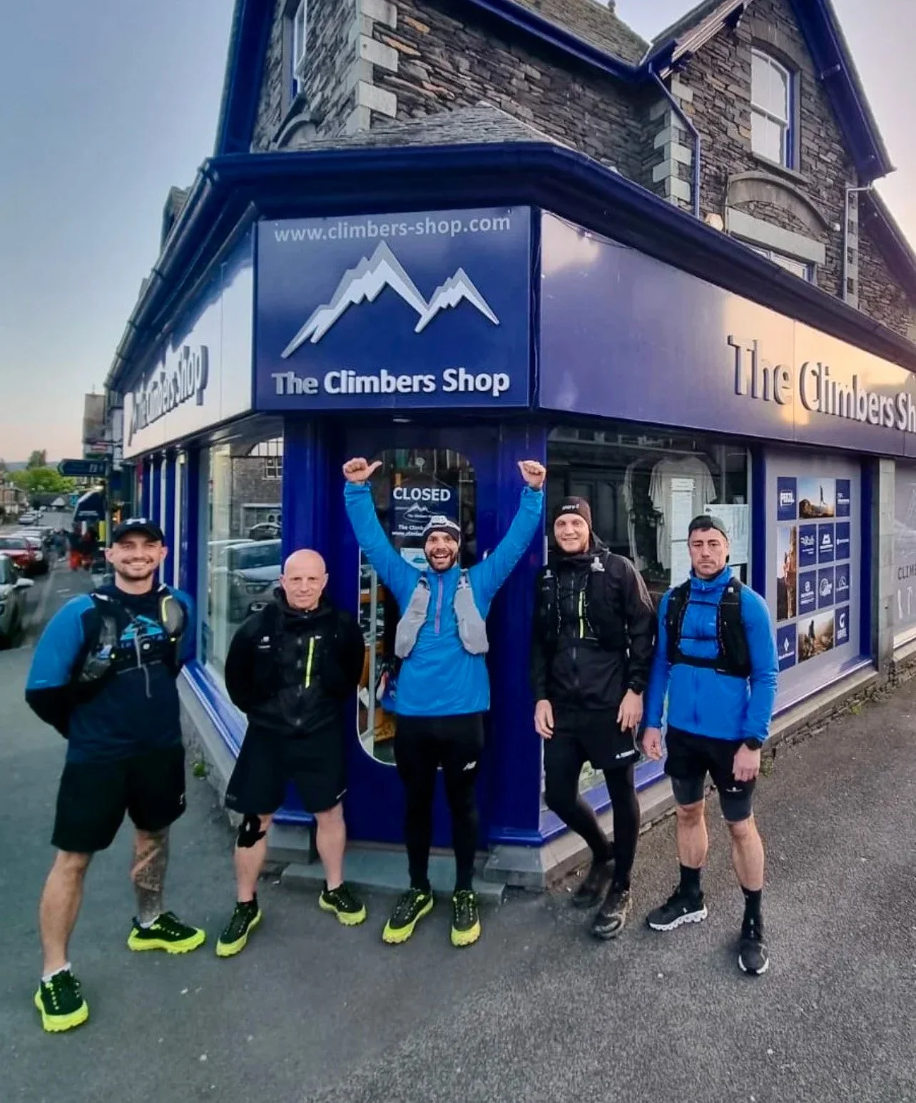 Five men wearing outdoor gear and backpacks standing outside The Climbers Shop, smiling, with one man raising his arms in celebration.