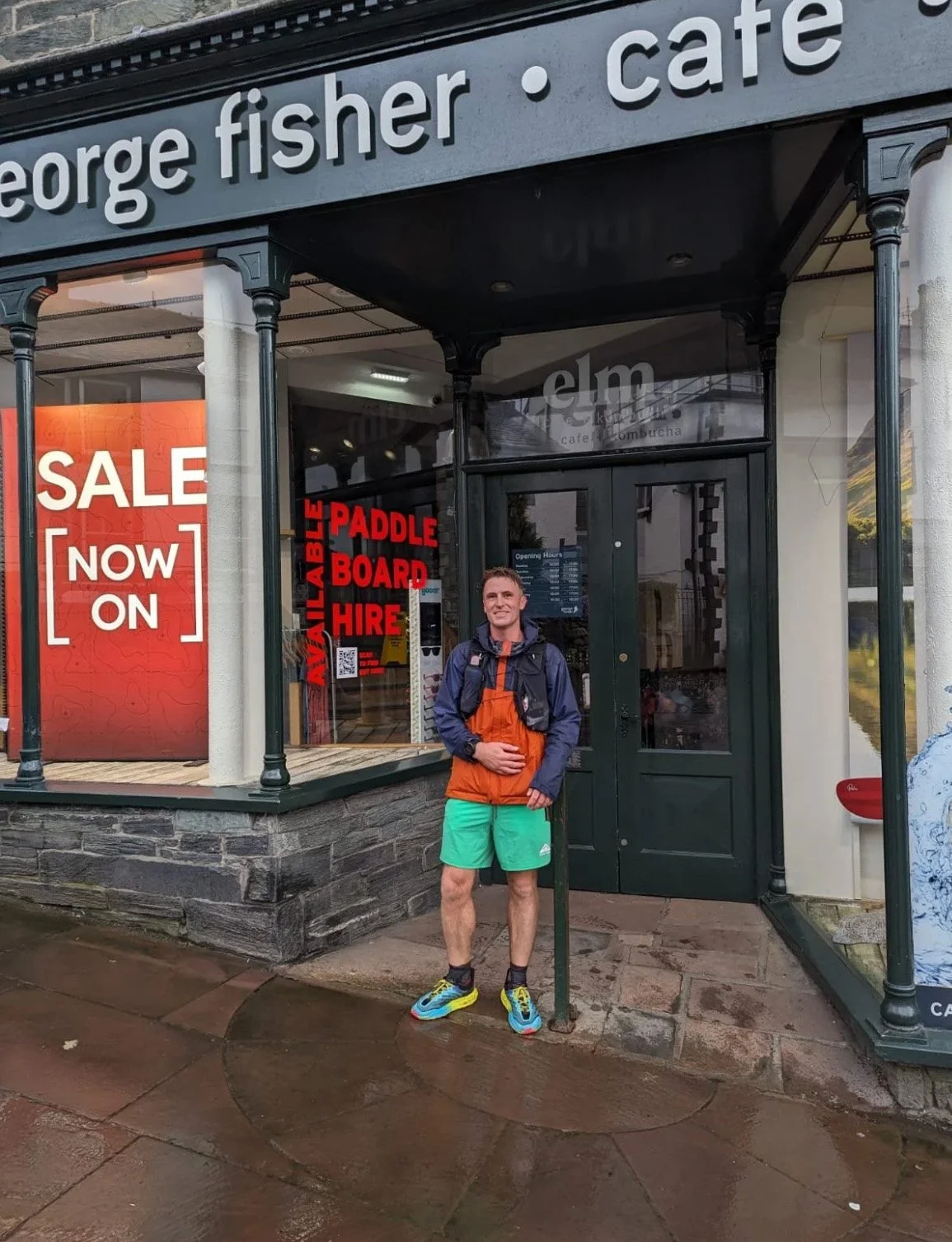 A man standing outside a store with a sign that reads 'sale now on' and another sign in the window advertising paddle board hire. The man is wearing a blue and orange jacket, green shorts, and colorful running shoes, smiling at the camera.