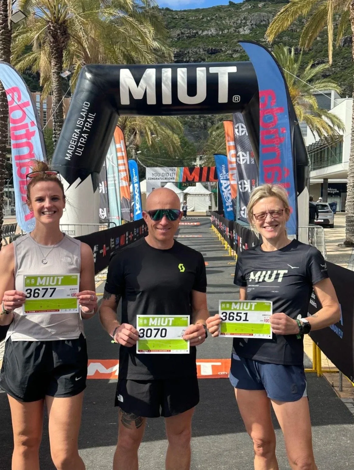 Three runners holding race bibs at the finish line of an outdoor ultra marathon event, with event branding and flags in the background.
