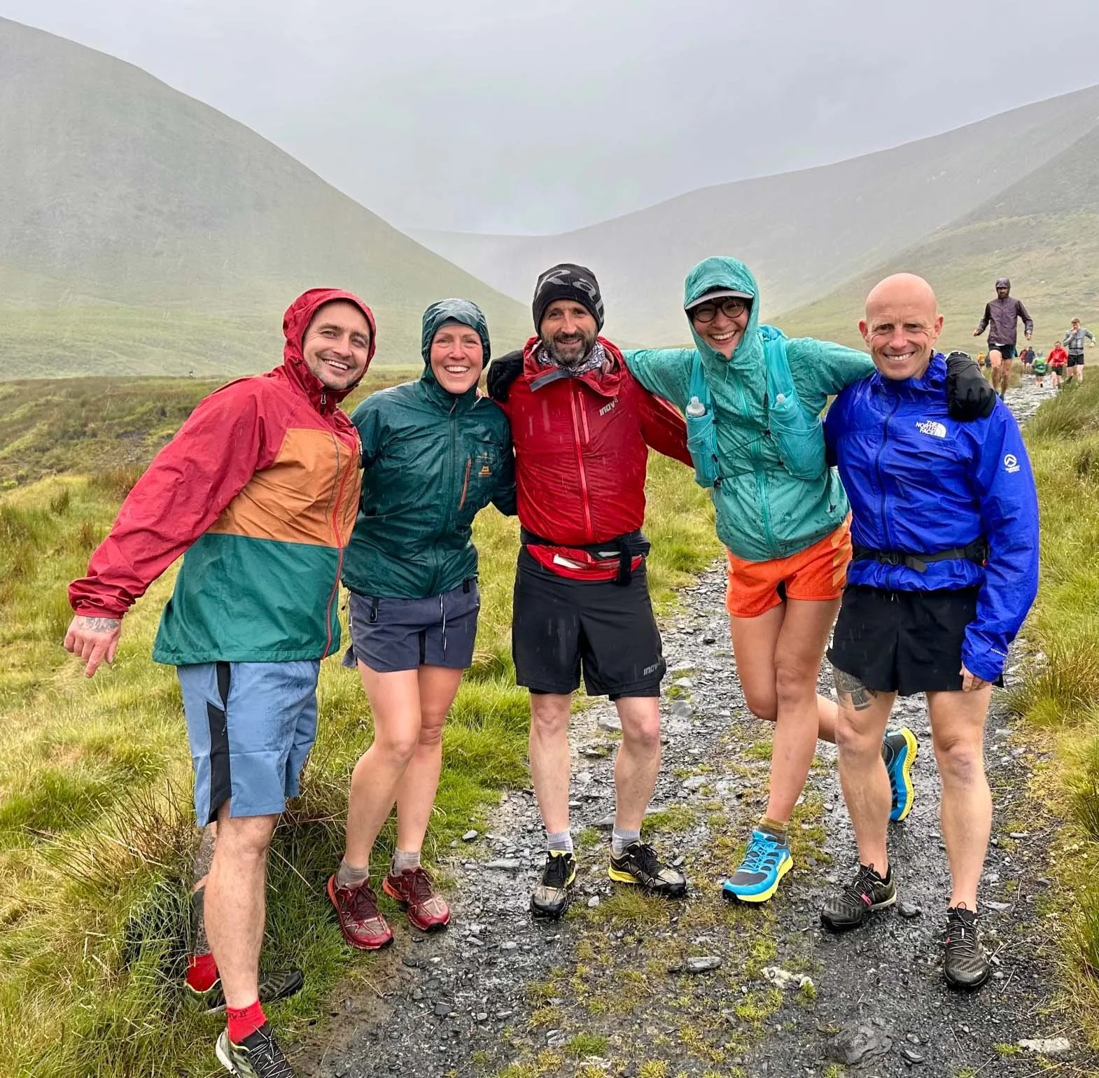 Group of five hikers dressed in rain gear, standing close together on a wet trail in a lush, green mountainous area, smiling at the camera.
