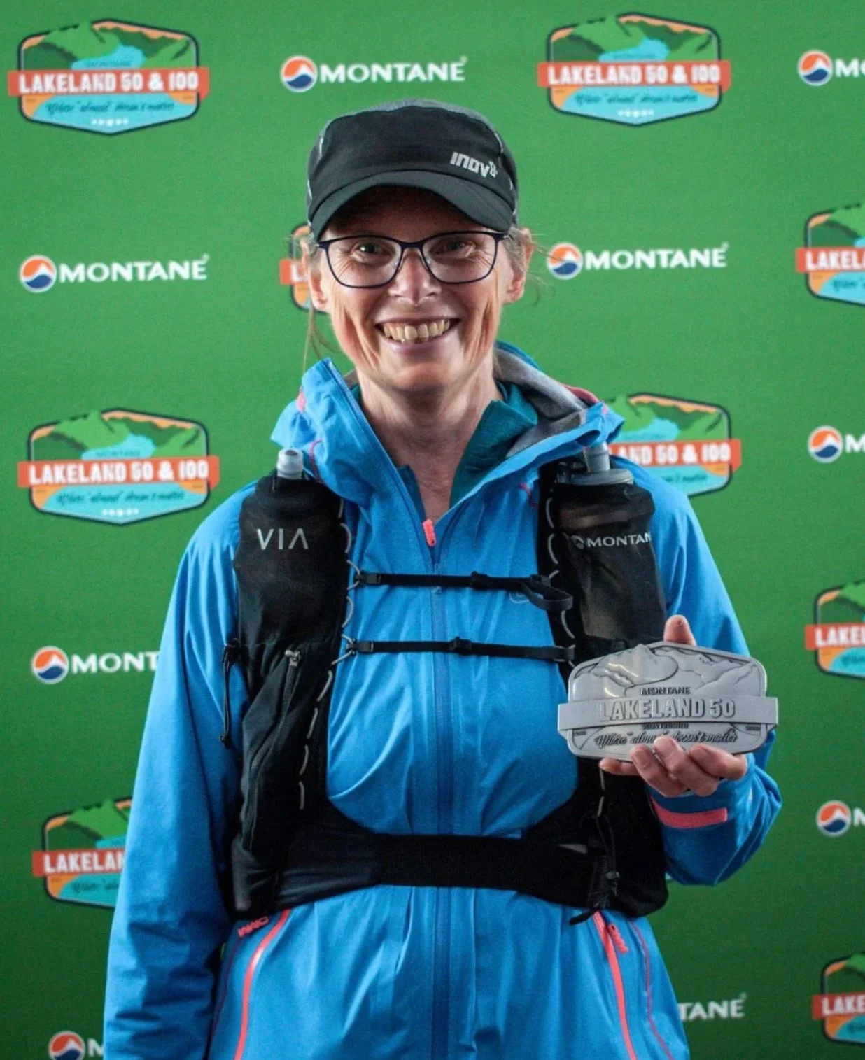 A smiling woman wearing a black cap, glasses, and a blue athletic jacket, holding a commemorative medal, standing in front of a green backdrop with logos for Montane and Lakeland 50 & 100.