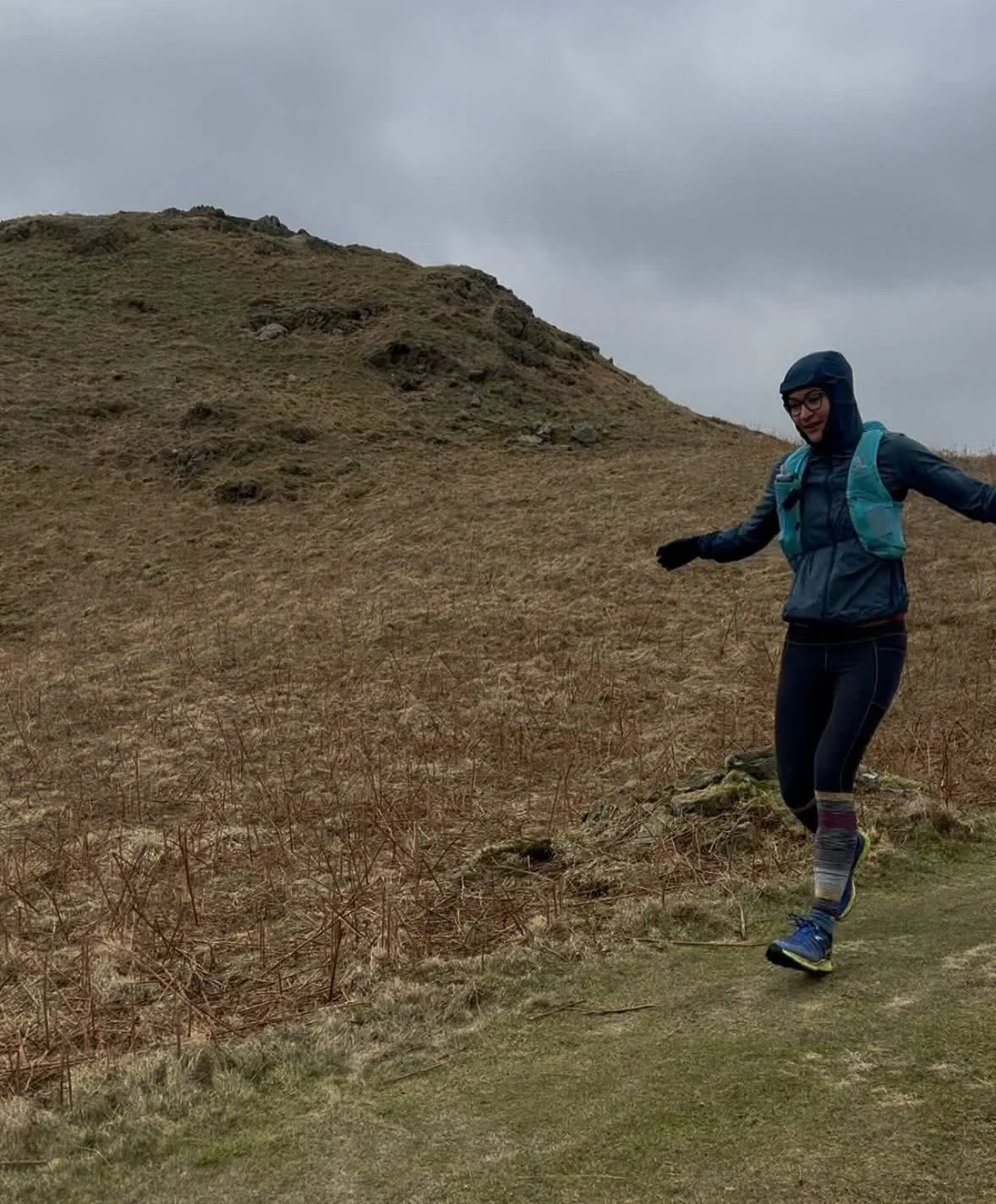 A person running on a mountain trail in outdoor gear, with hills and cloudy sky in the background.