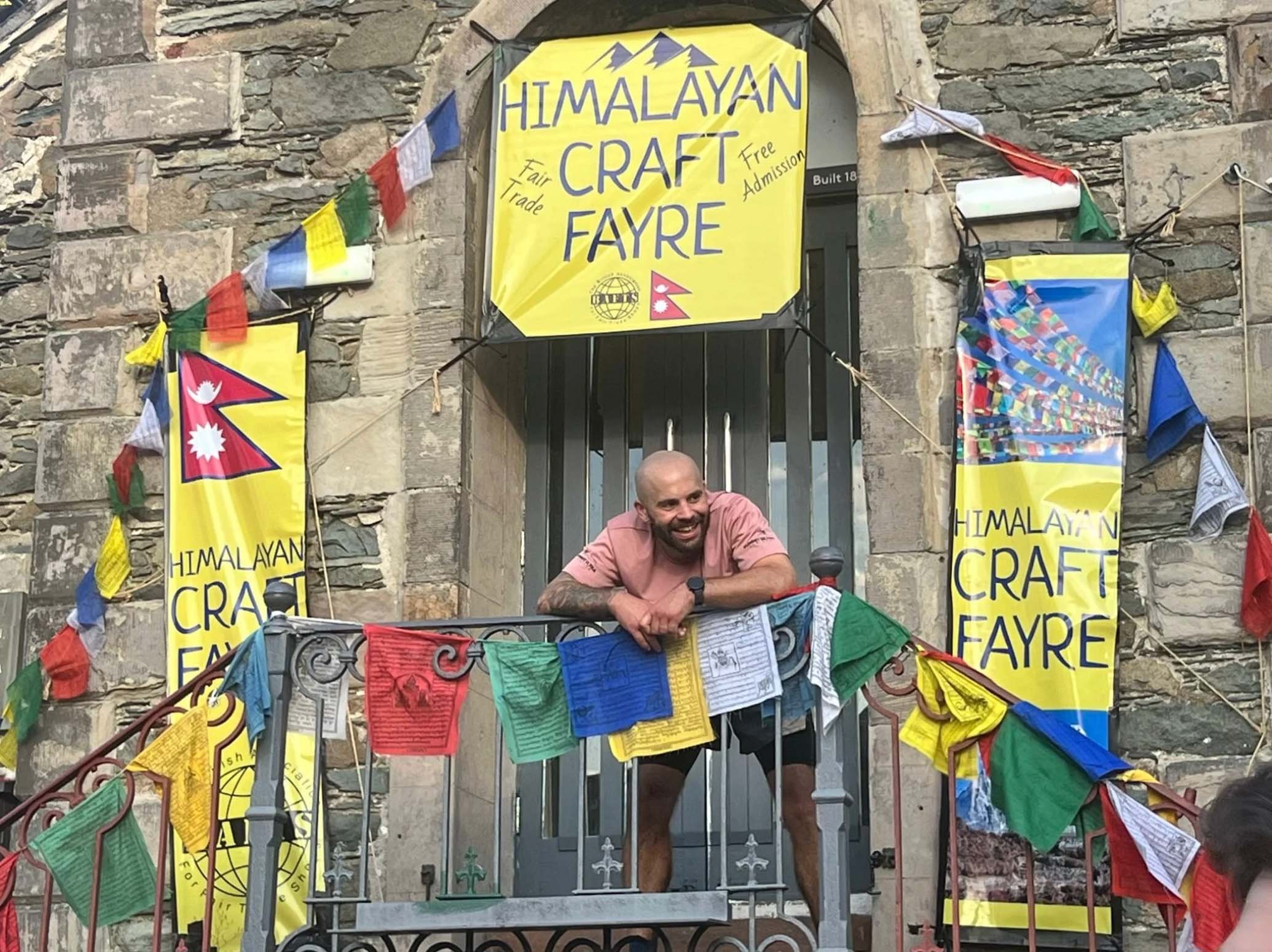 A man with a shaved head, beard, and tattooed arms smiling and leaning on a railing decorated with colorful prayer flags outside a stone building with Himalayan Craft Fayre banners.