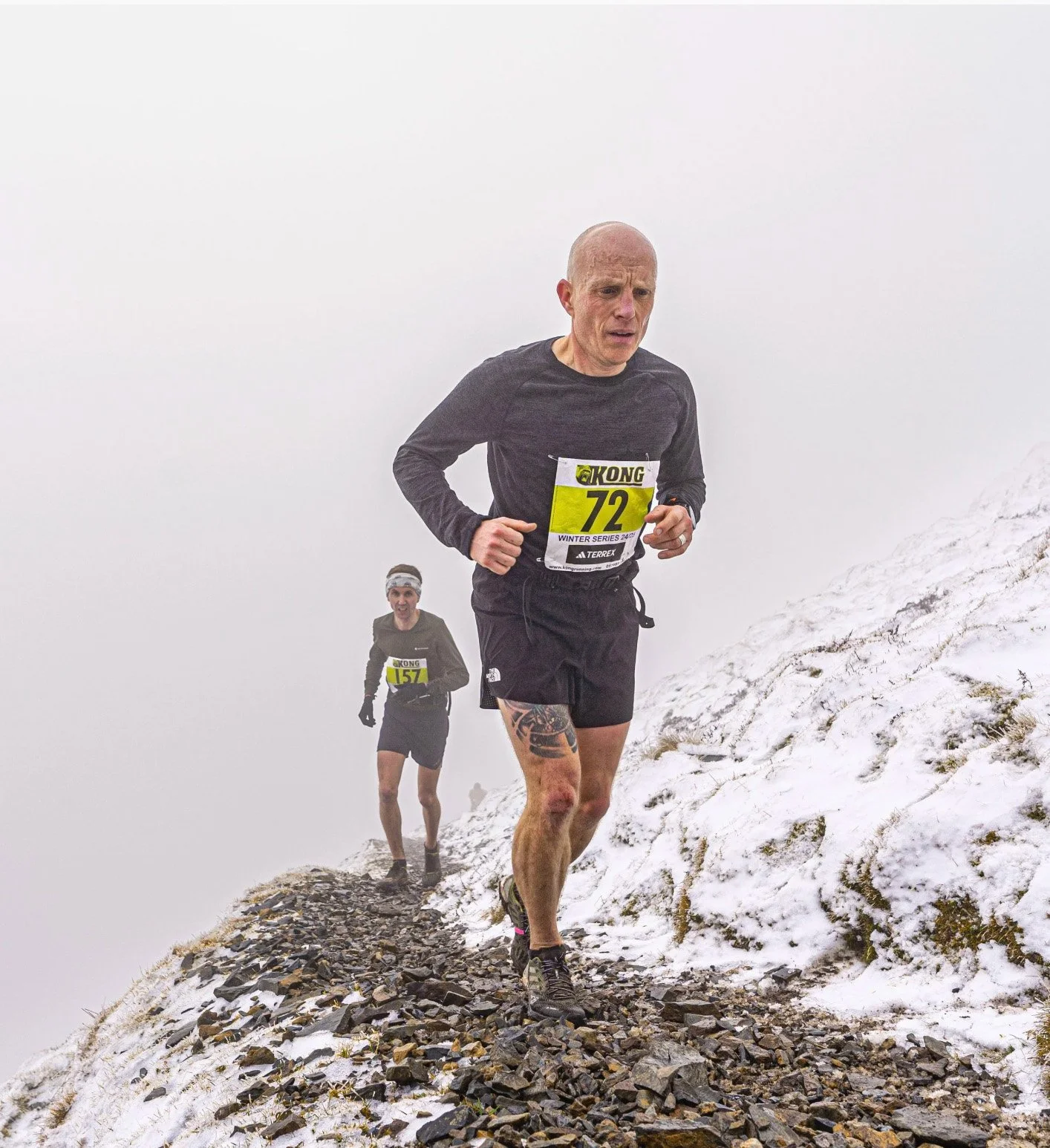 Two runners participating in a mountain race on a snow-covered trail. The front runner is a bald man wearing a black long-sleeve shirt, black shorts, and a yellow bib numbered 72. The second runner is a woman wearing a gray shirt, black shorts, and a headband, with a yellow bib numbered 157. The trail is rocky with patches of snow, and fog obscures the background.