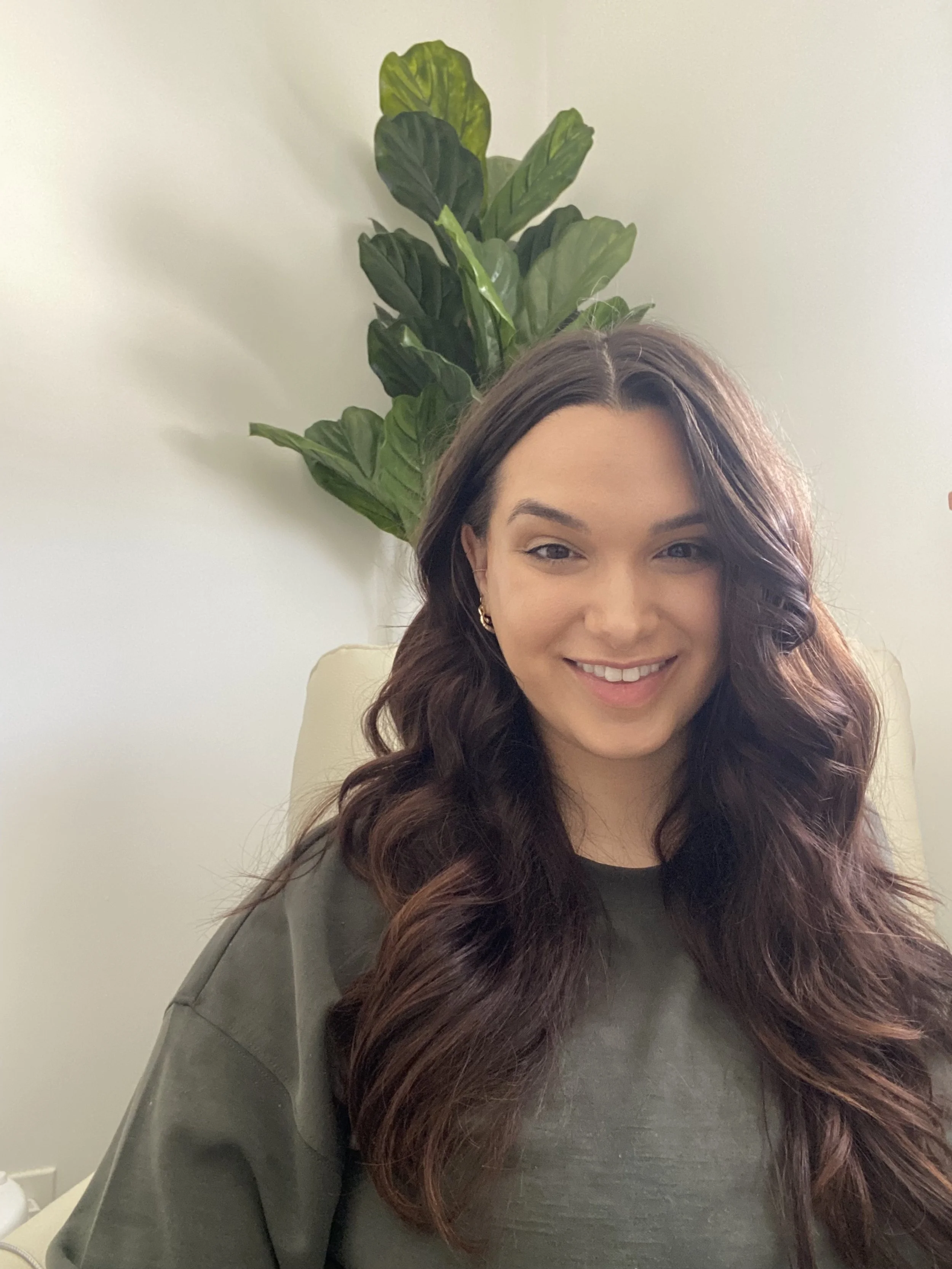 A woman with long wavy brown hair smiling while sitting in front of a green leafy plant and a light-colored wall.
