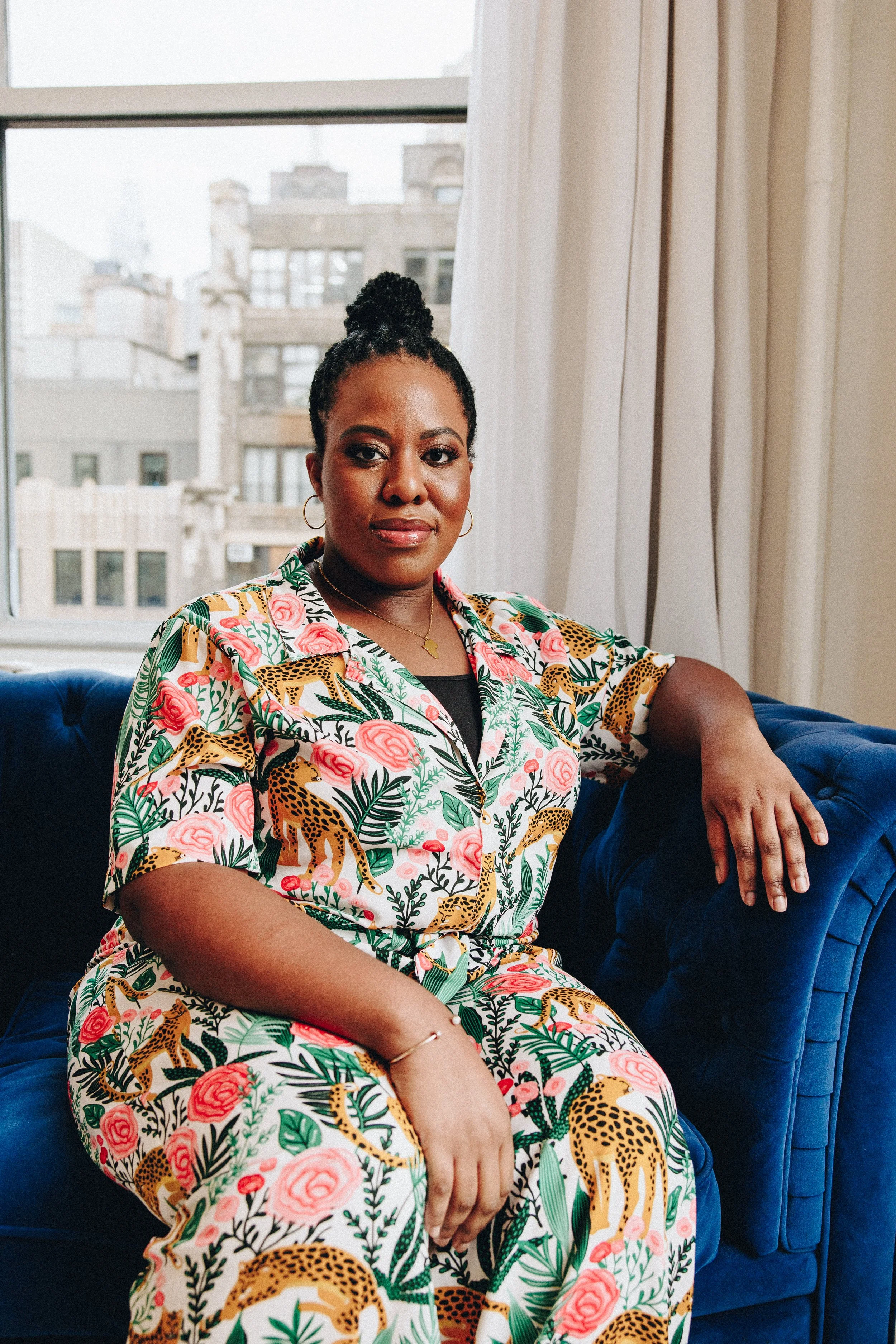 A dark-skinned Black woman sitting on a blue velvet sofa in front of a window with city buildings outside. She is wearing a colorful jumpsuit with leopards, pink roses, and green leaves, and has her arm resting on the sofa arm.