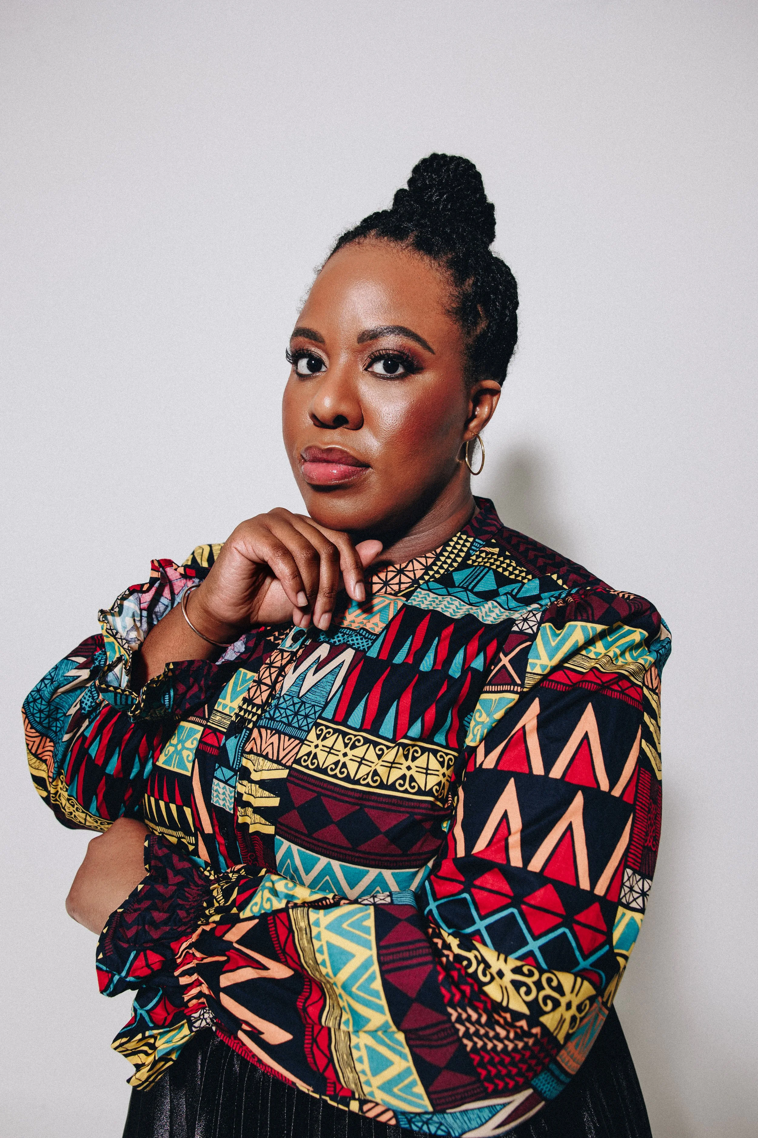 A dark-skinned Black woman with two-strand twists styled in a bun poses against a plain white background. She is wearing a colorful, patterned long-sleeve shirt with geometric designs in red, yellow, blue, and black.