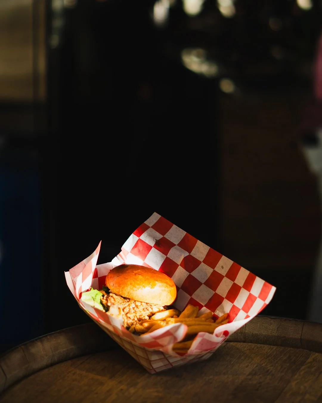 A serving of smash burger with fries on red-and-white checkered paper, placed on a dark wooden surface, in a dimly lit setting.