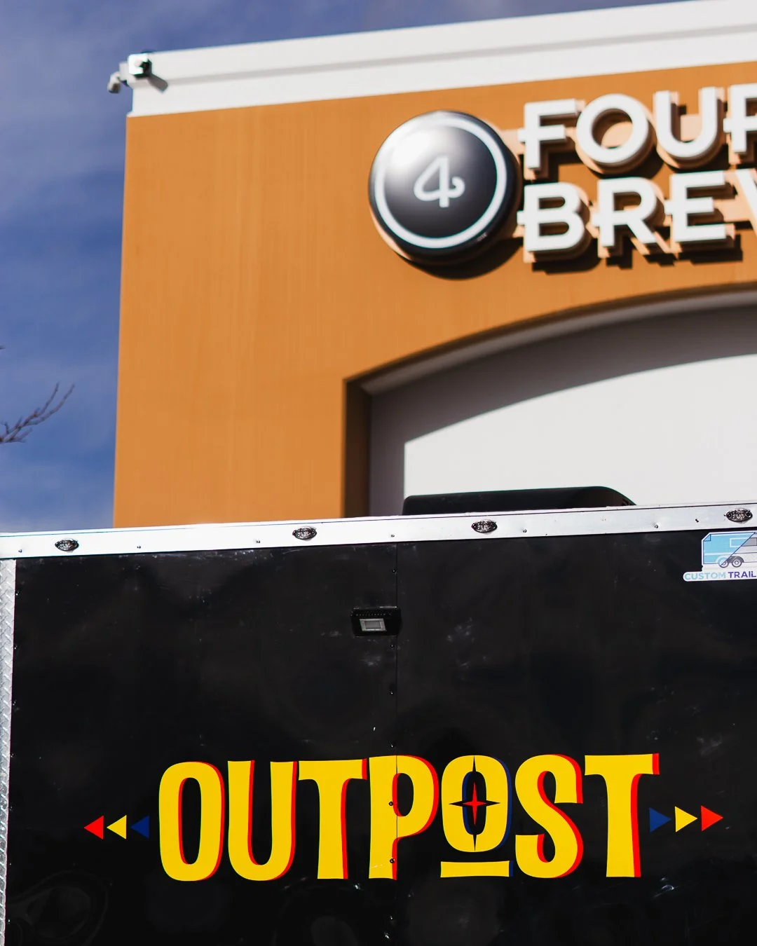 A sign for 4 Noses Brewing Company with the number 4 inside a circle, and a black trailer with the word OUTPOST in large yellow and red letters.