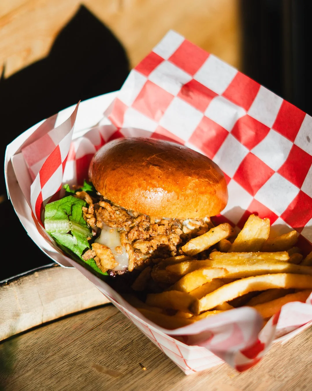 A burger with lettuce, ground meat patty, melted cheese, and a shiny bun, served with French fries in a red and white checkered paper liner.