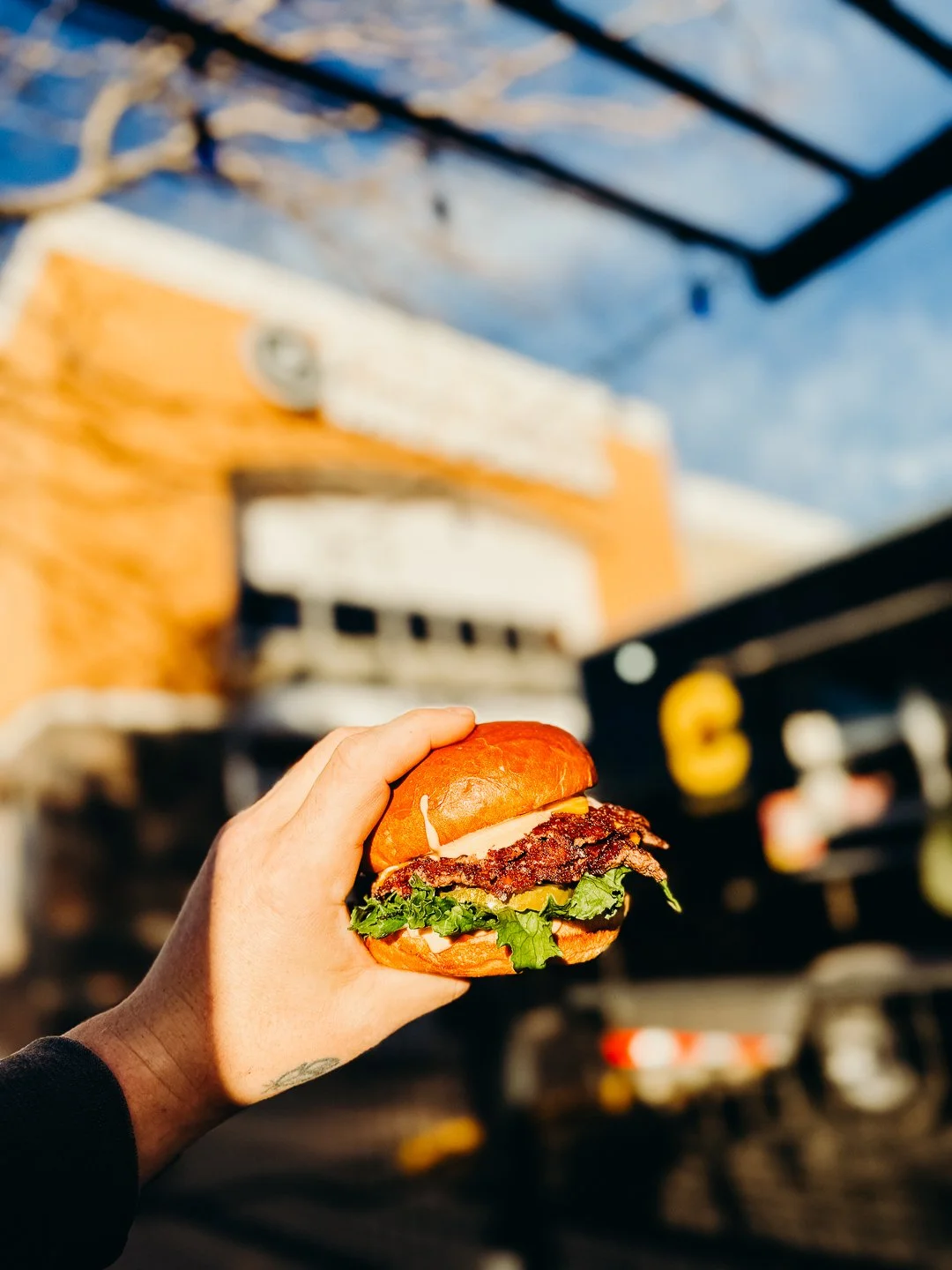 Person holding a burger with lettuce, tomato, and a beef patty outside near a food truck.