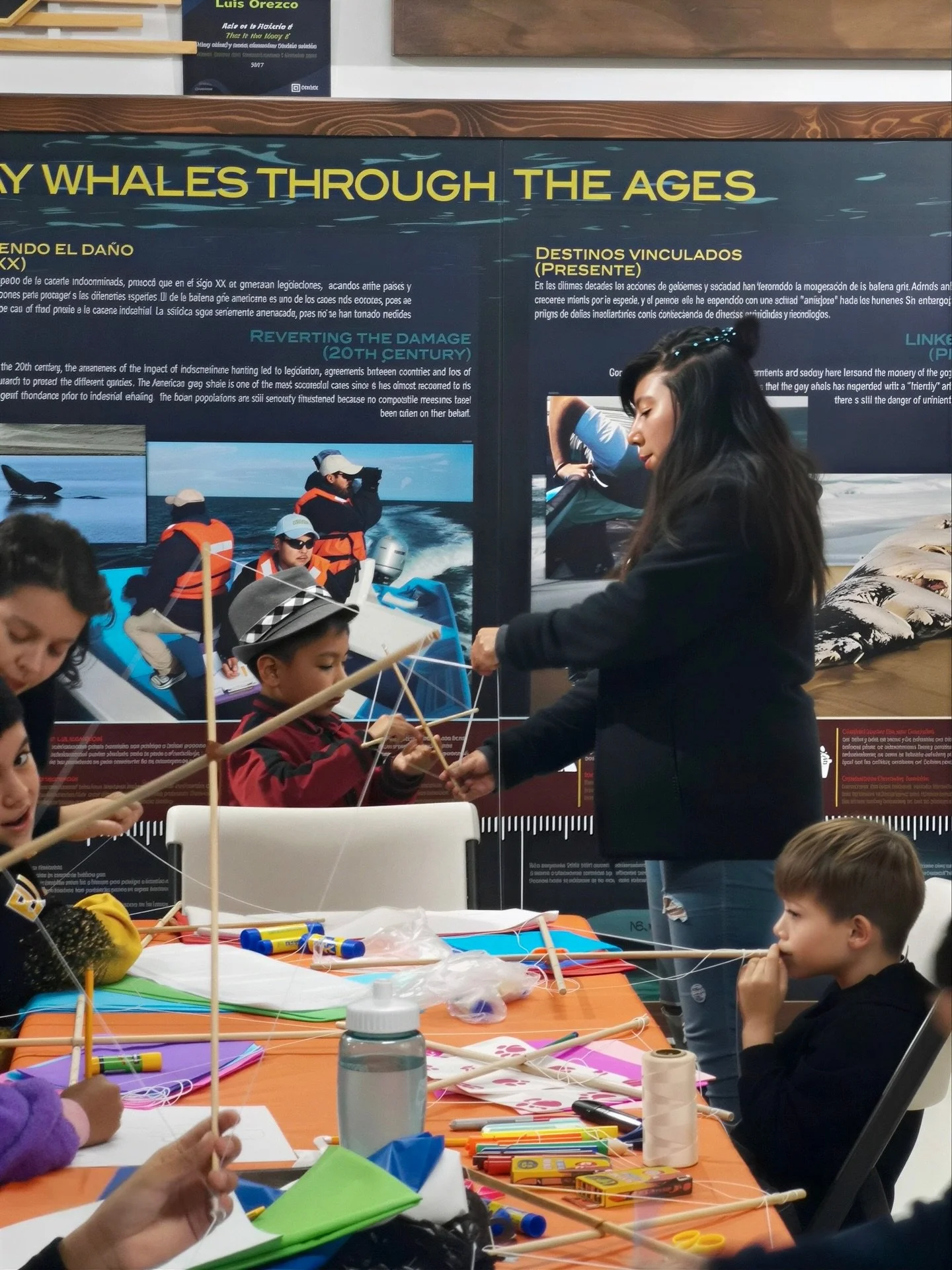 Niños y adultos creando modelos de ballenas con materiales de manualidades en una mesa en un entorno educativo. En el fondo hay un cartel con información sobre ballenas y el océano.