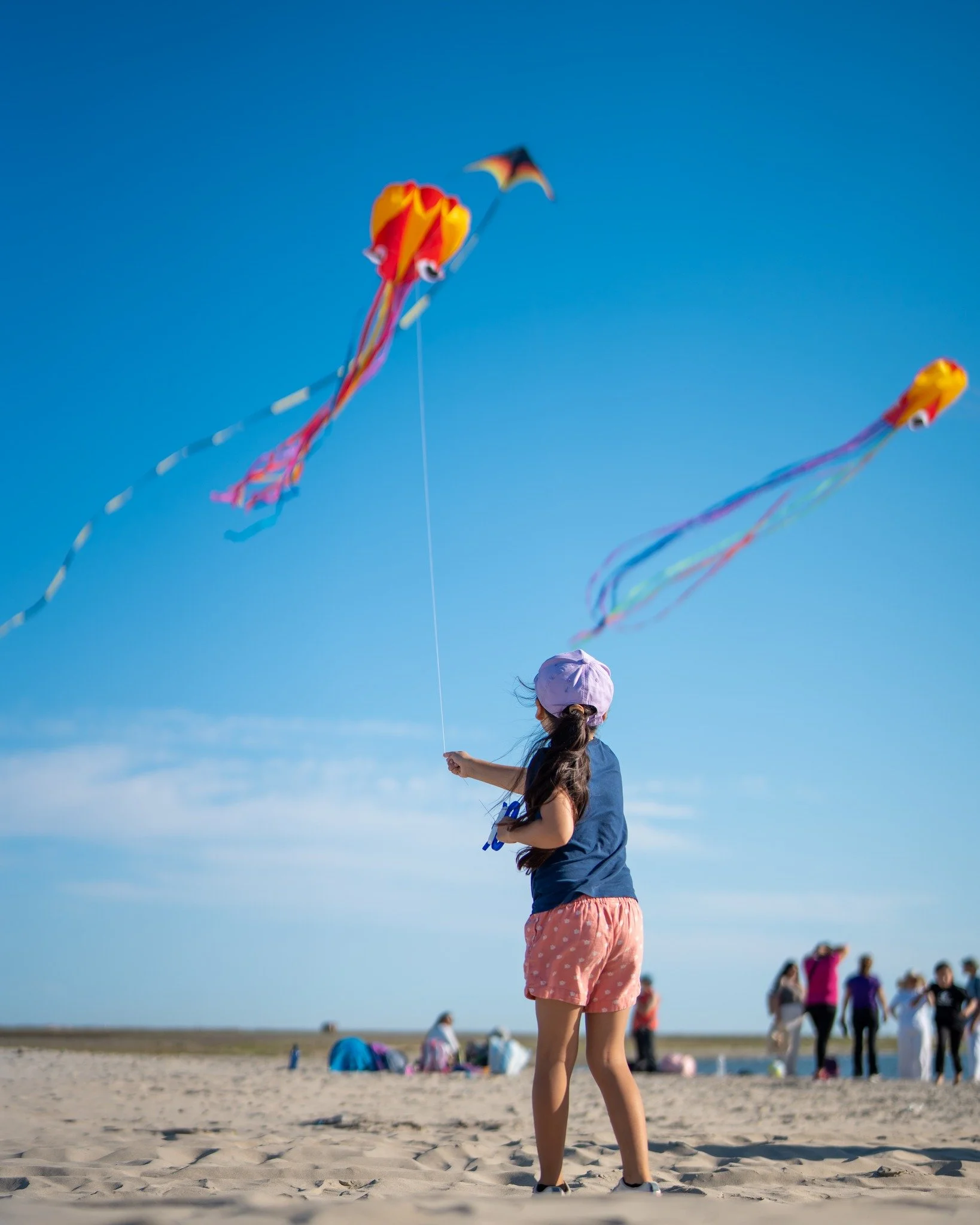 Niña en la playa con cometas de colores en el cielo azul, otras personas en el fondo, algunos con bolsas y ropa informal, en un día soleado.