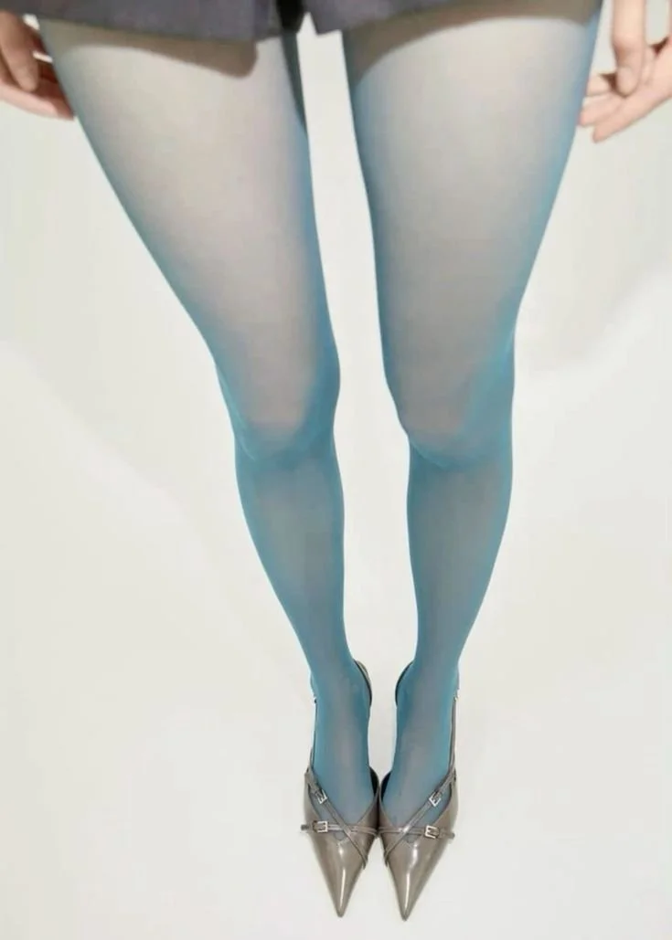 Close-up of a person's legs in sheer blue tights, wearing pointed high heels, with the person standing on a white background.