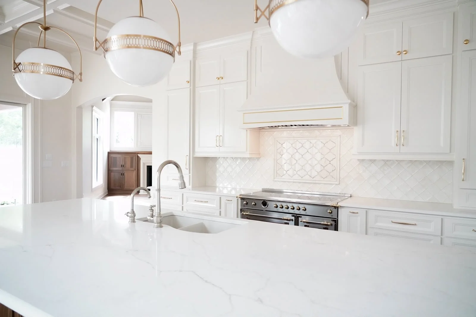 Clean, white kitchen with marble countertops, gold cabinet handles, and a stainless steel oven, illuminated by natural light.