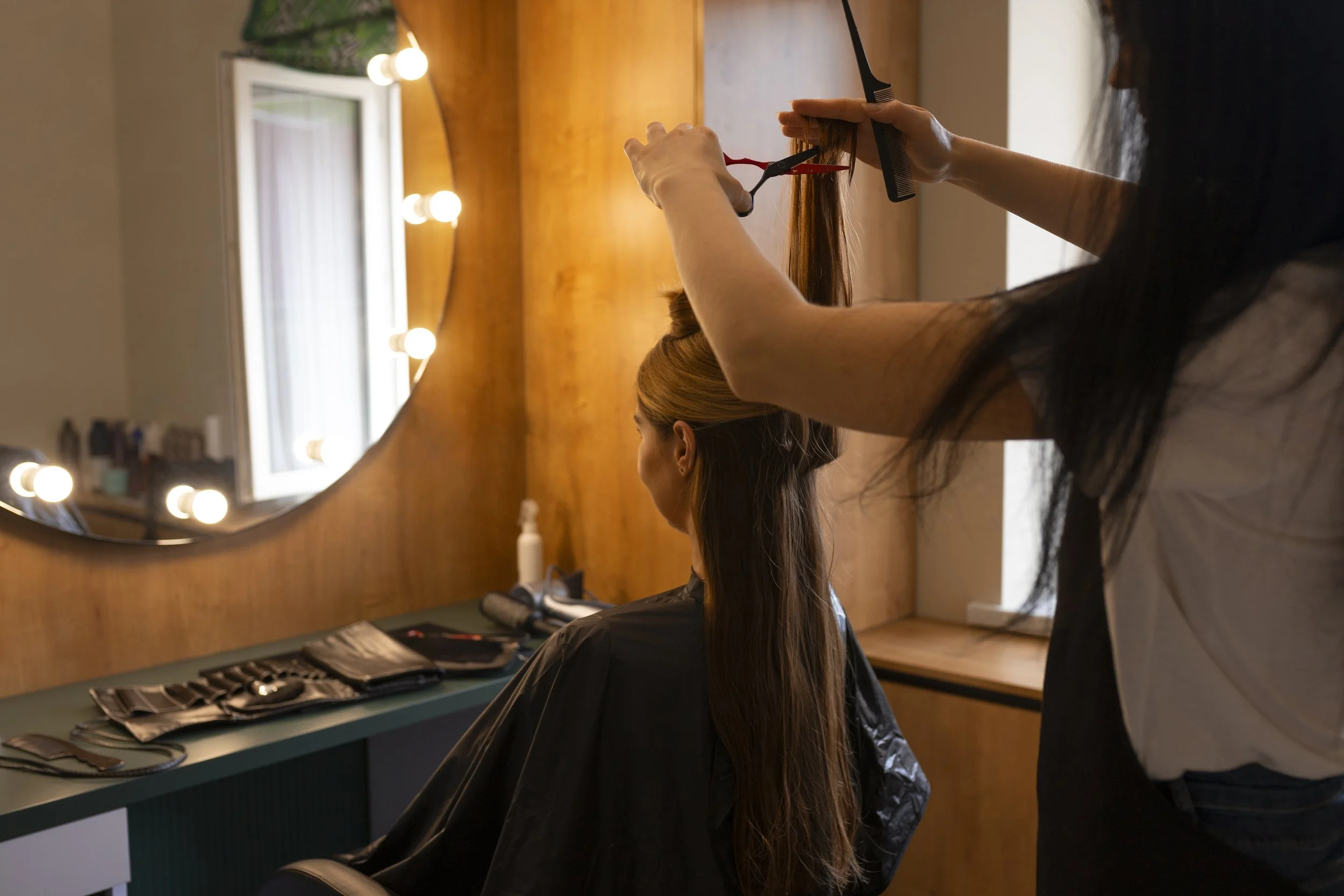Hair stylist cutting a woman's long brown hair in a salon with a large mirror and warm lighting.