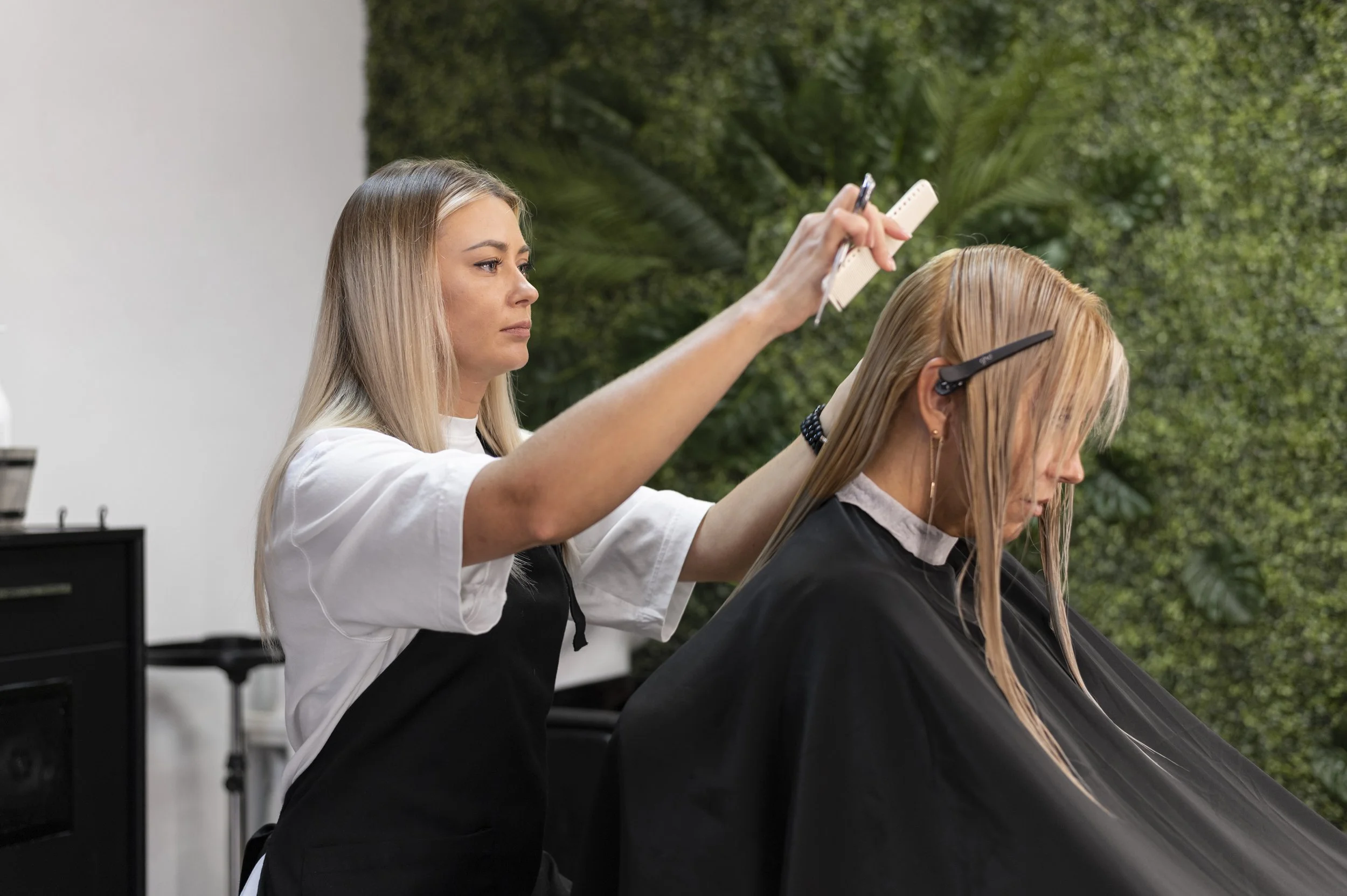 A hairstylist cutting a client's hair in a salon with a green foliage background.