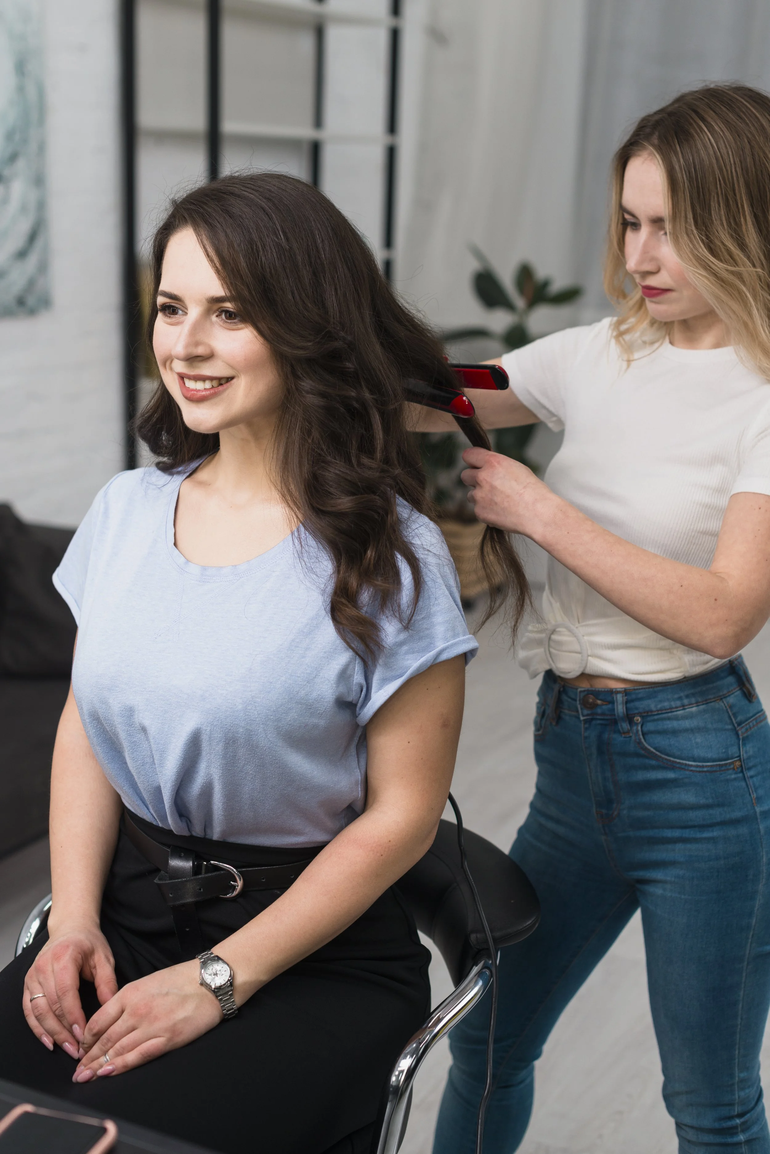 A woman with long, dark, wavy hair is getting her hair styled by a hairstylist in a salon. The woman is smiling and seated in a chair, wearing a light blue T-shirt and a watch on her left wrist. The hairstylist is using a flat iron to curl her hair.
