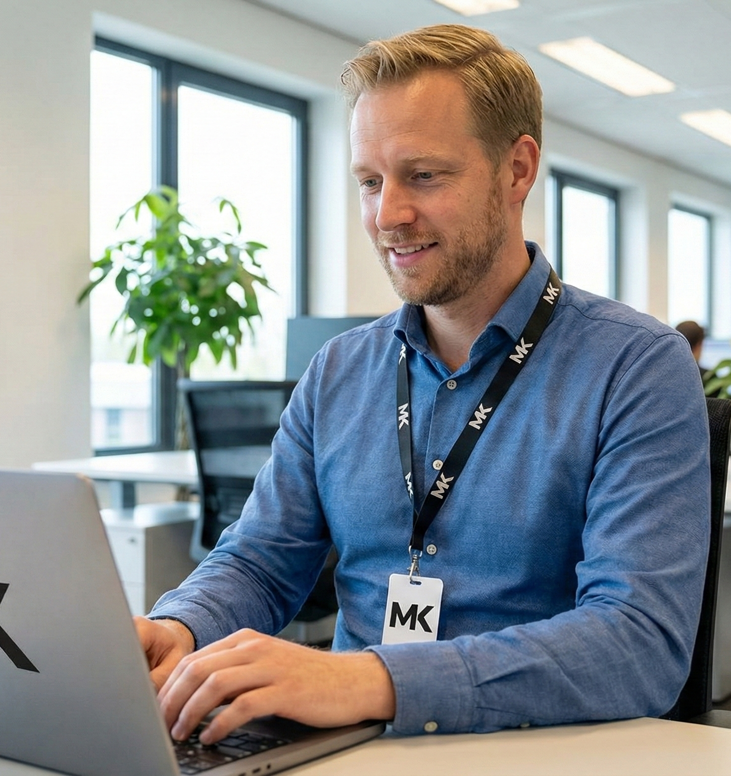 Man die in een kantoor werkt, zittend voor een laptop, met een MK badge om zijn nek, glimlachend.