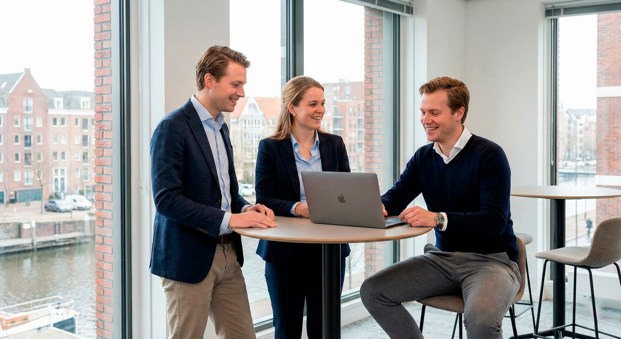 Drie zakenvrouwen en zakemannen die een gesprek voeren rond een tafel met een open laptop, in een modern kantoorgebouw met grote ramen en uitzicht op de stad.