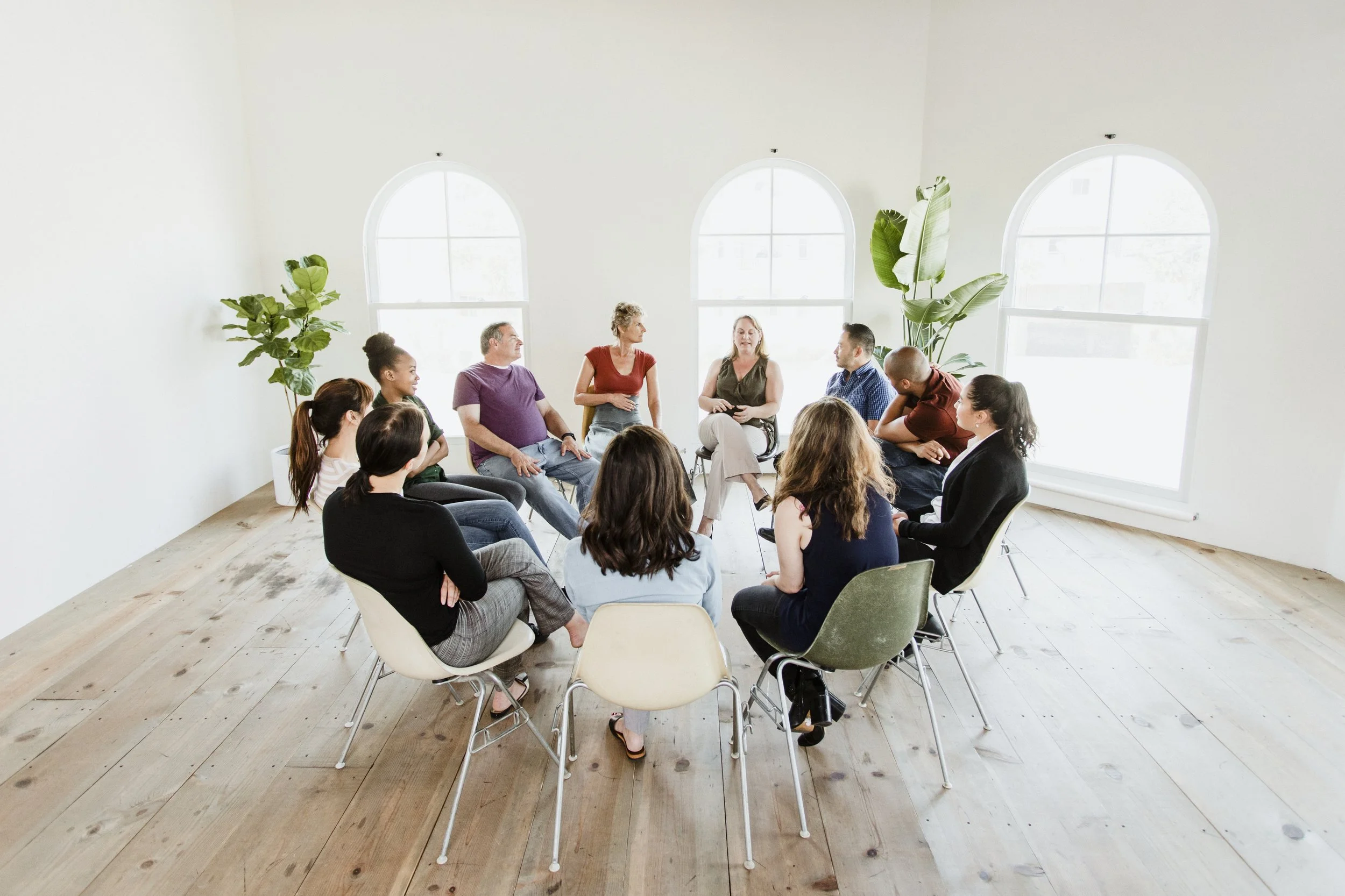 A group of people sitting in a circle during a discussion or therapy session, in a bright room with large arched windows and potted plants.