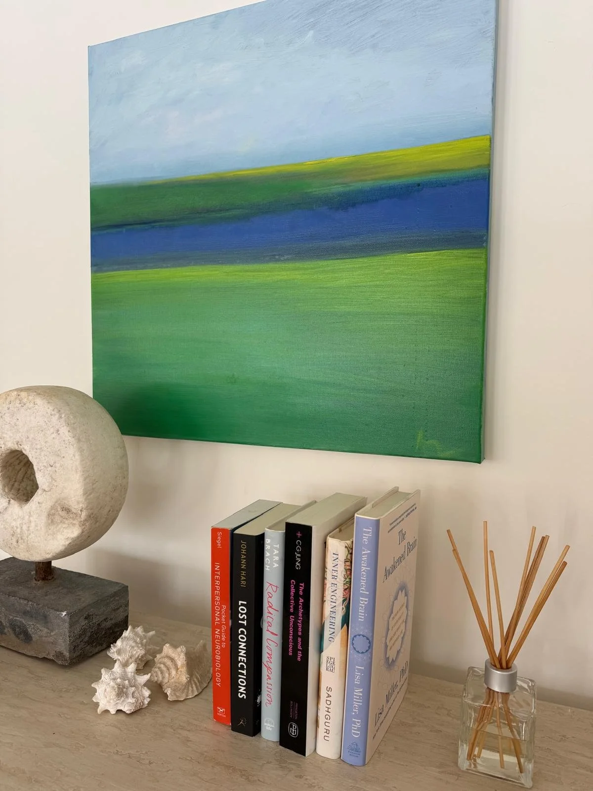 A collection of books on a wooden surface next to decorative items, including a stone sculpture, seashells, and a reed diffuser, with a colorful landscape painting by Ana Jardim Messenger hanging on the wall behind.