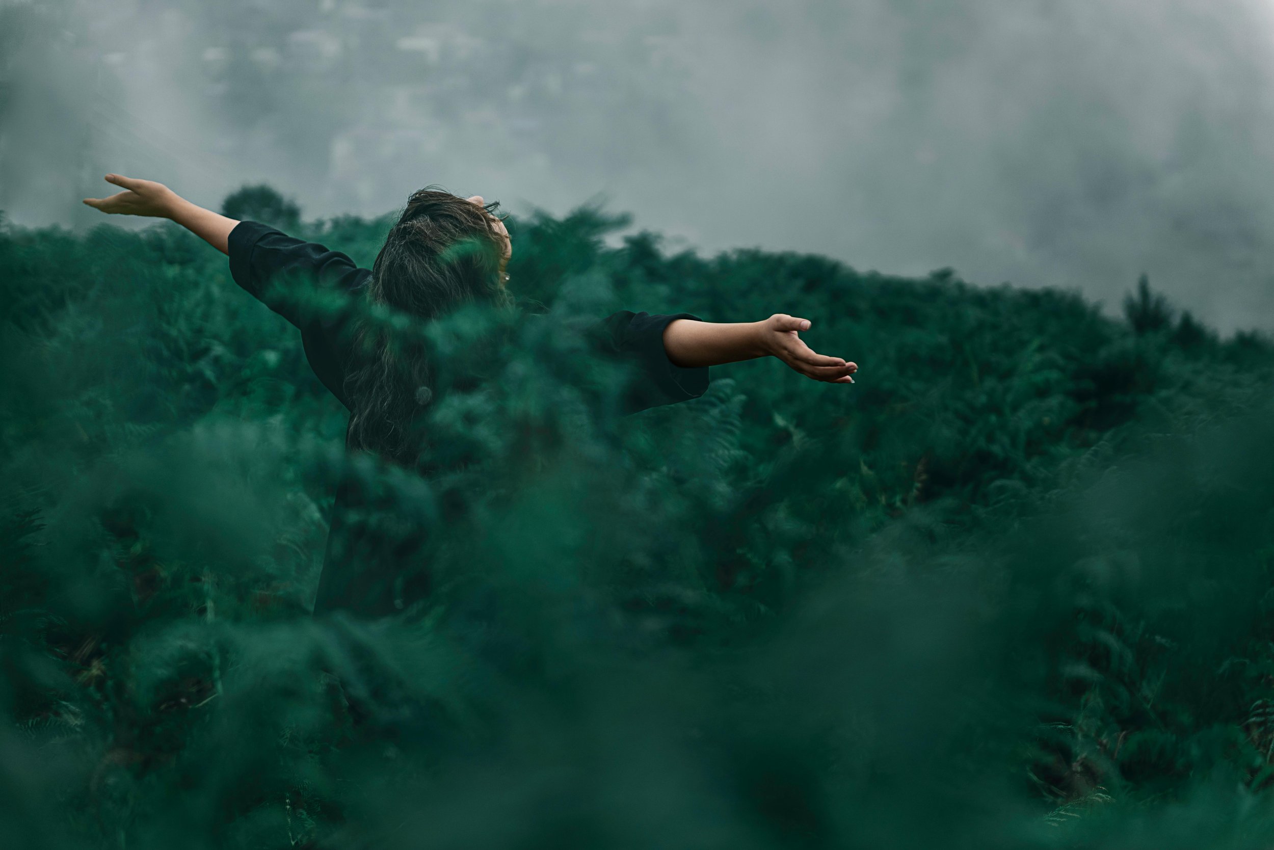 A person with long hair and wearing a black top is standing outdoors with arms extended to the sides, against a background of lush green trees and cloudy sky.