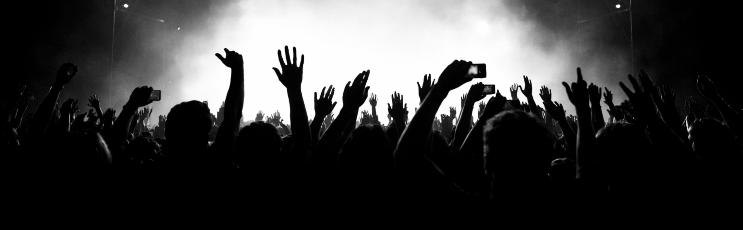 Silhouettes of concertgoers with raised hands, some holding smartphones, in front of a brightly lit stage at a concert.