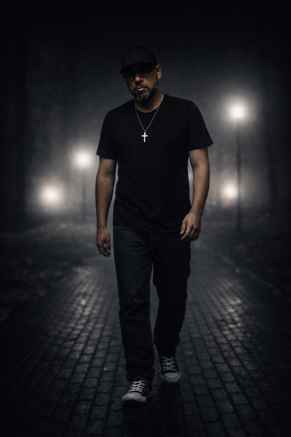 A man walking on a dark, wet street at night, wearing glasses, a baseball cap, a black t-shirt, jeans, sneakers, and a silver cross necklace, with streetlights in the background.