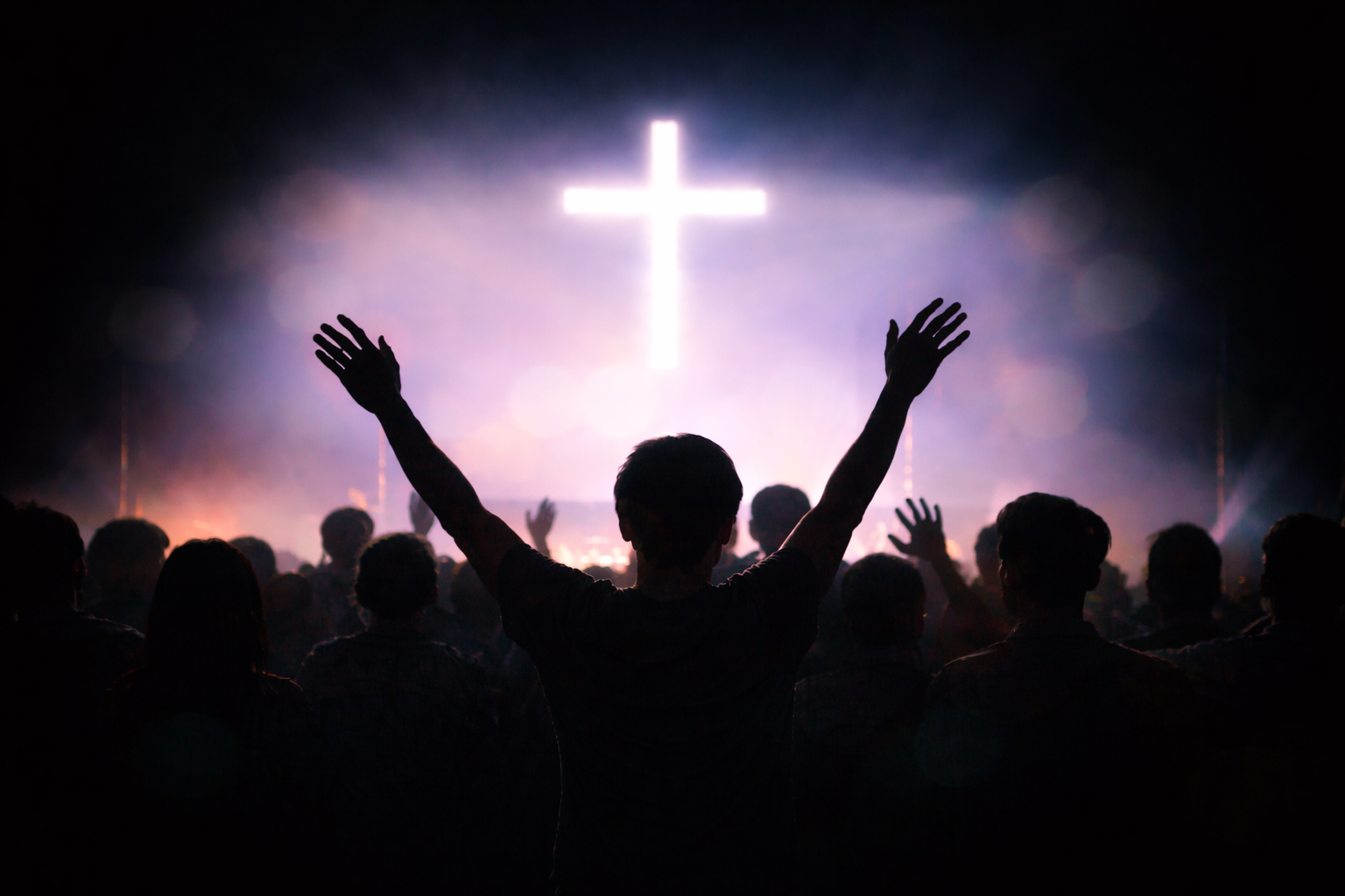 Person with raised hands in front of a large illuminated cross, crowd gathered indoors or at an event.