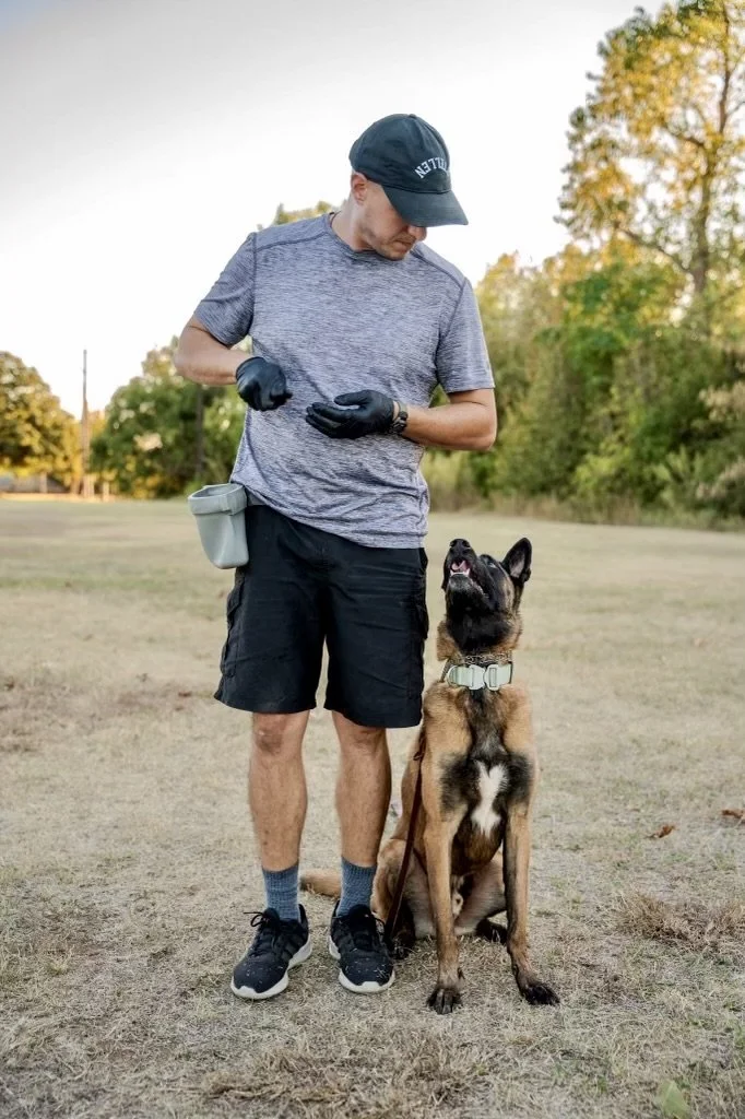 Man wearing black shorts, a grey t-shirt, a black cap, black gloves, and black shoes standing outdoors with a Belgian Malinois dog sitting attentively by his side. The man appears to be training or giving commands to the dog in a park during fall.