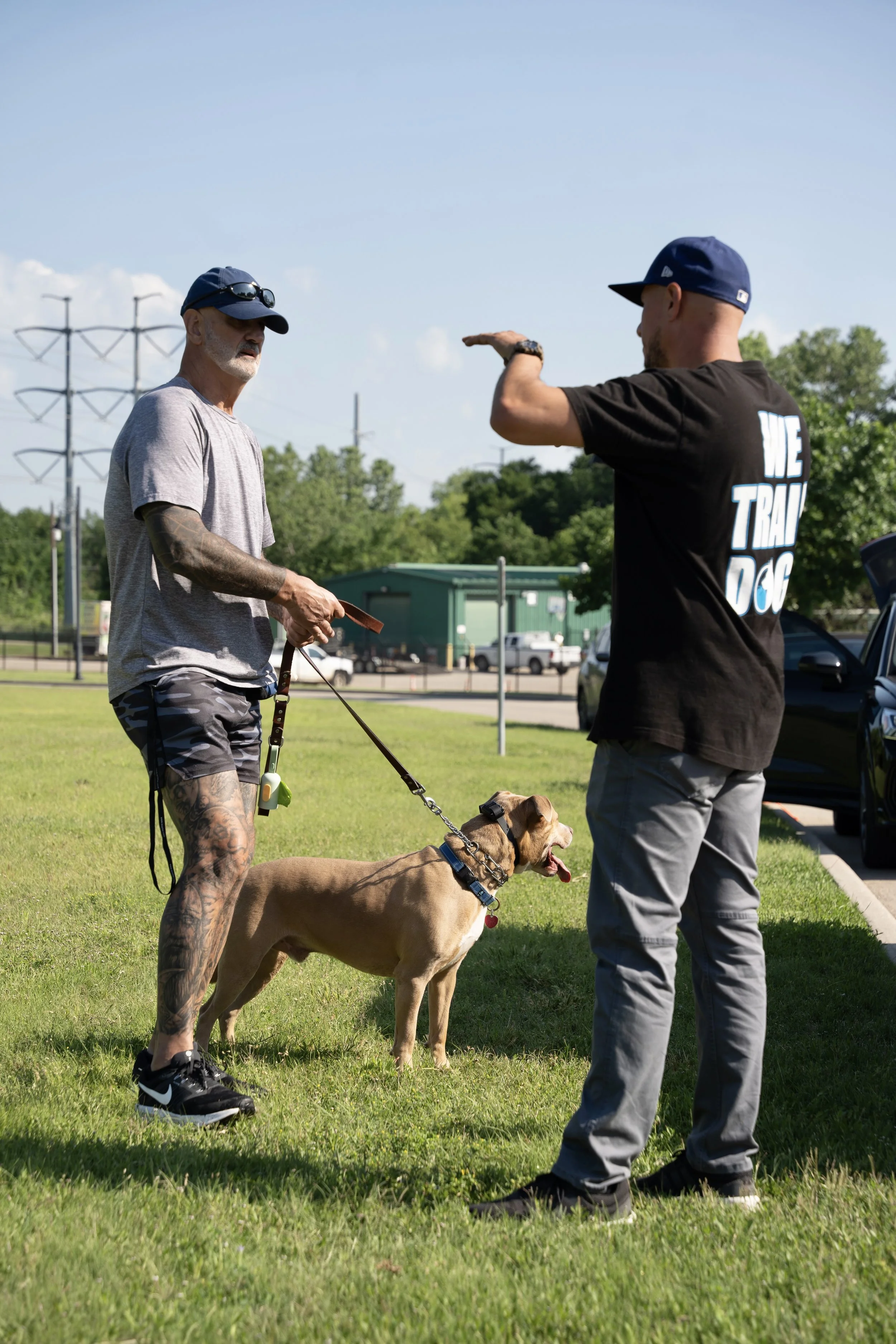 Two men talking outdoors on a grassy area with a dog between them. One man has tattoos, sunglasses, a cap, and shorts, holding a leash attached to the dog. The other man is wearing a black shirt with white text on the back that says 'WE TRAIN DOGS,' and a cap. The dog is a brown and black dog with a mouth open, panting.