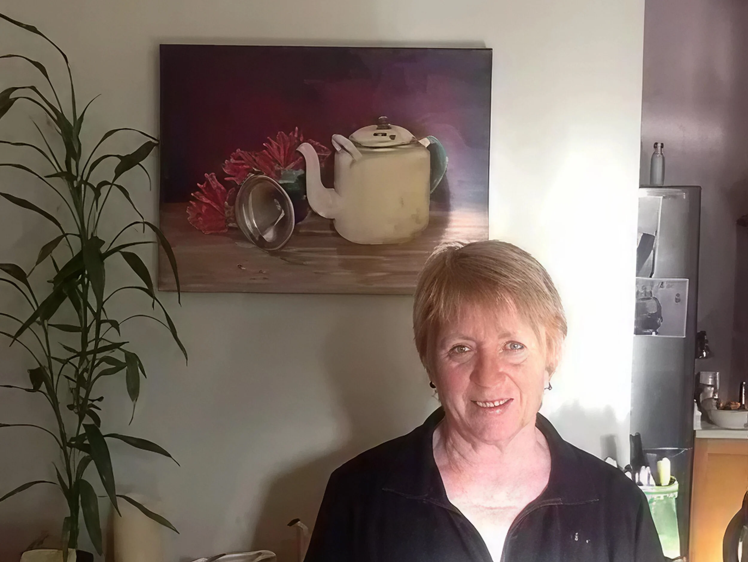 A smiling woman with short blonde hair standing in a kitchen near a tall leafy plant, with a painting of a teapot and flowers on the wall behind her, and a stainless steel appliance to her right.