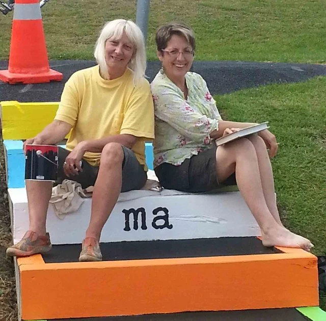 Two women sitting on a large colorful bench with the partially visible letters 'ma' on it, outdoors on a grassy area with traffic cones nearby. One woman has white hair and is wearing a yellow shirt, and the other has short brown hair and is wearing a floral blouse. They are smiling and appear to be enjoying a casual moment.