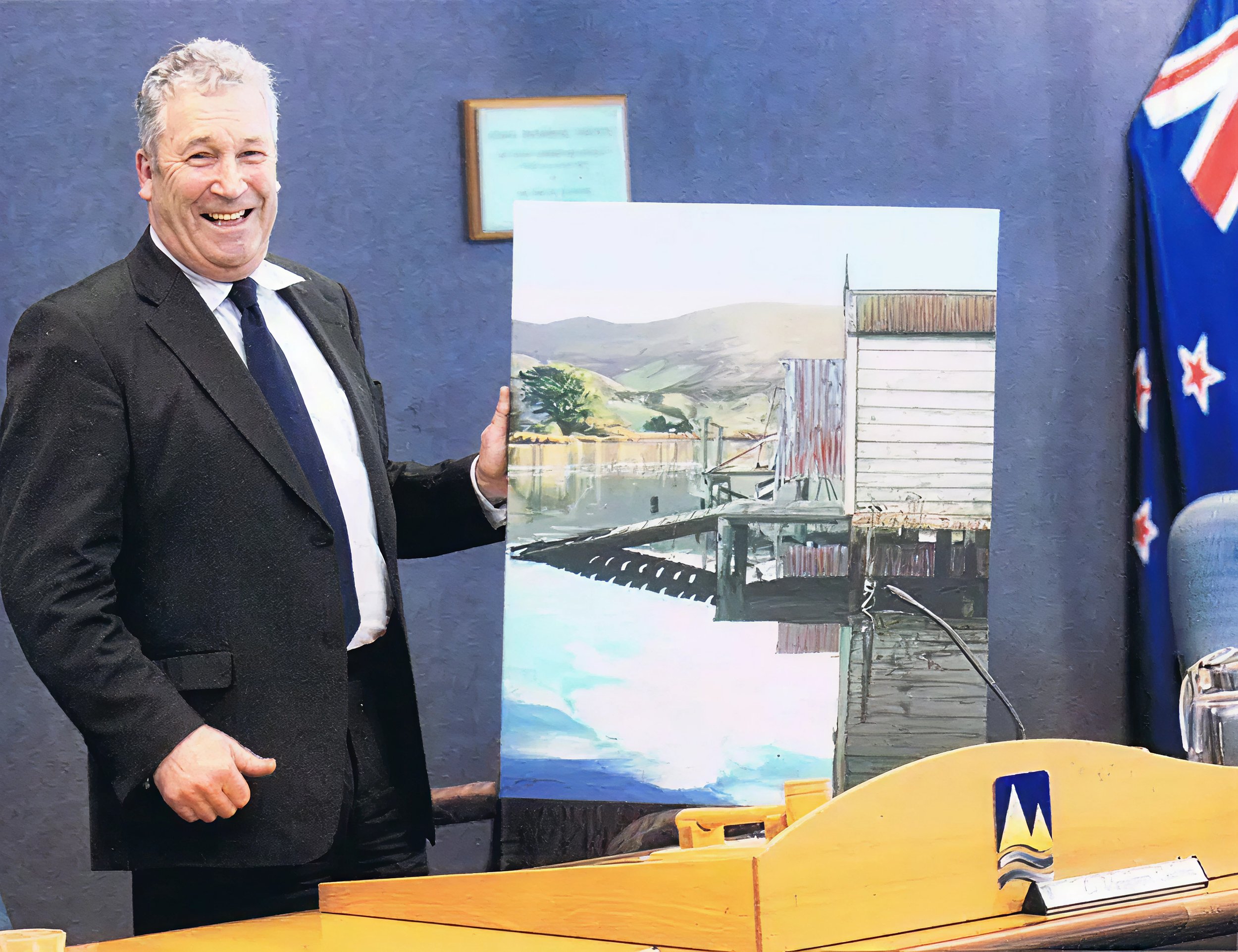 A man in a black suit and tie is smiling and holding a painting of a waterfront scene, with a dock, buildings, and mountains in the background. He stands beside a yellow table in a room with blue walls, with an Australian flag visible on the right side.