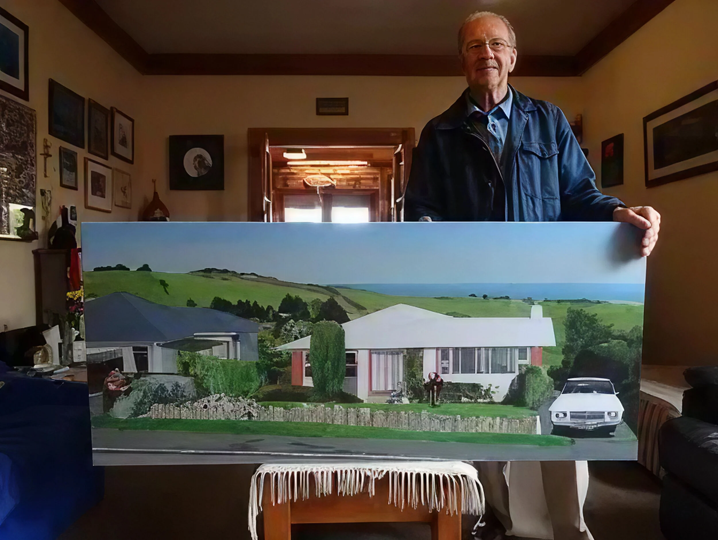 A man standing indoors holding a large painting of a house with a white roof, surrounded by green hills and a car in the driveway.